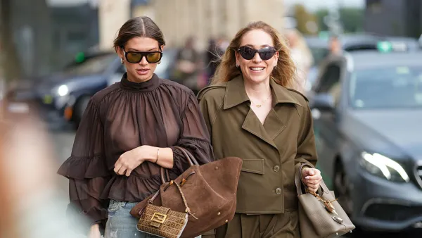 COPENHAGEN, DENMARK - AUGUST 06: A guest wears a sheer, dark brown blouse with a high neckline and layered ruffle sleeves. The hair is styled in a sleek, center-parted updo. Sunglasses are oversized with a rectangular frame. Earrings are small and circular. A bracelet is worn on the wrist. The jeans are light blue with a distressed finish. A large brown suede bag is carried, featuring gold hardware. A smaller brown Fendi monogrammed bag with a top handle is also visible. Another guest wears sunglasses with dark lenses and a brown frame. The hair is styled in loose waves. A brown double-breasted jacket features a notched lapel, button closure, and a flap pocket on one side. The sleeves are rolled up. Matching high-waisted trousers have a pleated front and a relaxed fit. A taupe textured handbag is carried, with short handles, outside Gestuz, during Copenhagen Fashion Week, on August 06, 2025 in Copenhagen, Denmark (Photo by Edward Berthelot/Getty Images)