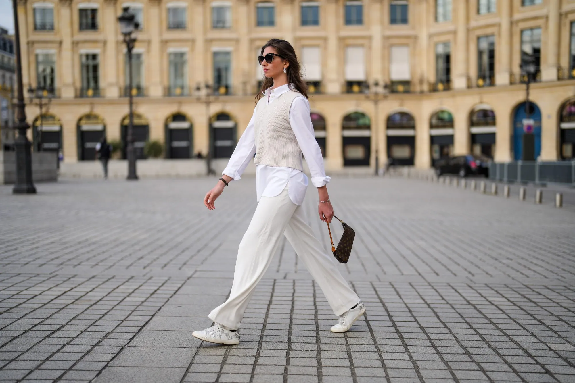 PARIS, FRANCE - APRIL 30: Amanda Derhy @amanda_jashley wears black sunglasses, pearls earrings, an oversized shirt, large flared pants, a beige sleeveless ribs wool pullover, white and silver leather Zadig & Voltaire laces sneakers, silver bracelets, a brown coated canvas monogram Louis Vuitton Lucky Me bracelet, a brown monogram in coated canvas Louis Vuitton Pochette Accessoires shoulder bag, on April 30, 2021 in Paris, France. (Photo by Edward Berthelot/Getty Images)
