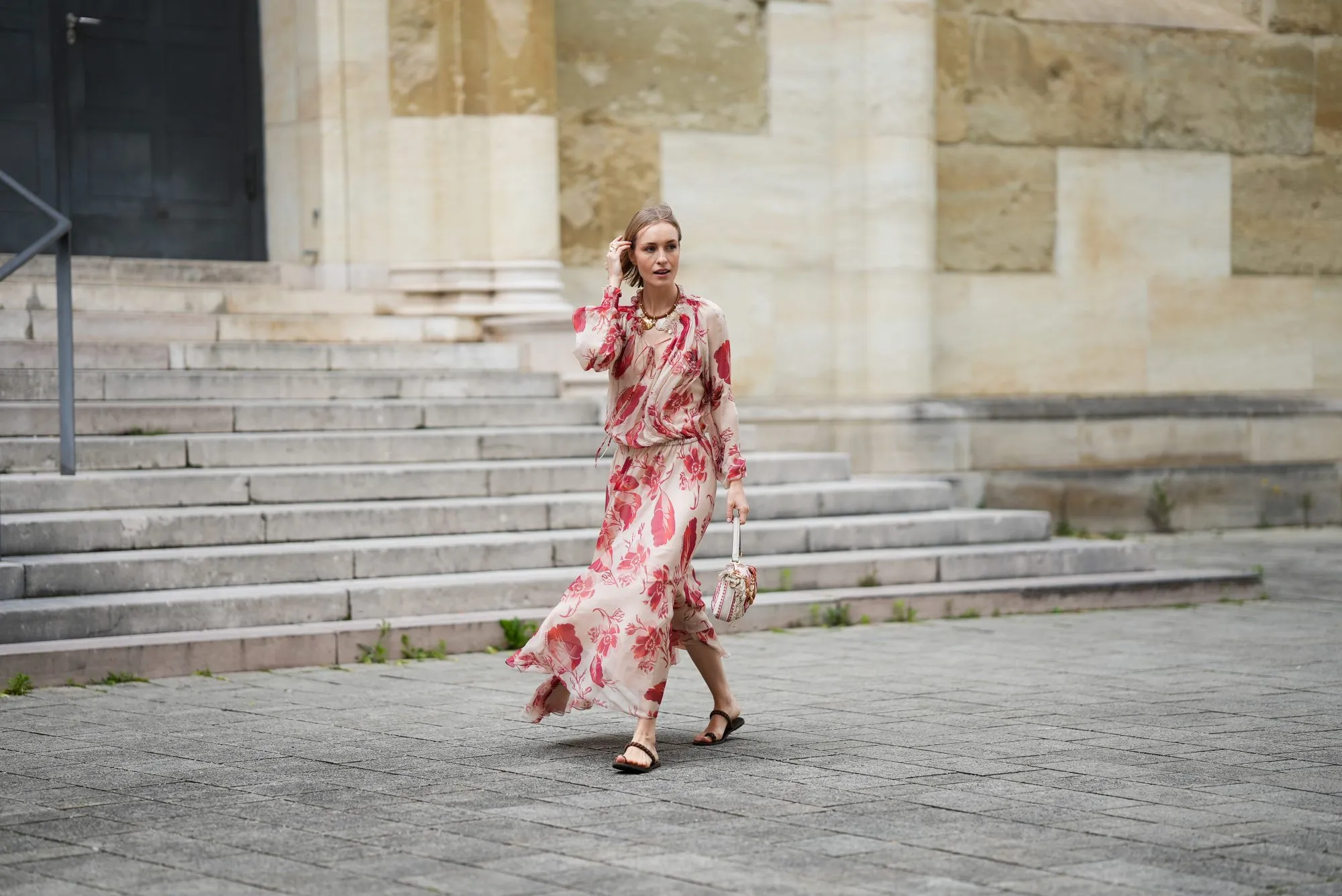 MUNICH, GERMANY - MAY 29: Marlies Pia Pfeifhofer is seen wearing a light beige silk chiffon long sleeve maxi dress with red and pink prints of feathers and flowers, drawstring waist, deep V-neck with front tie and smocking details on the collar and cuffs from FENDI, a natural-colored Baguette canvas bag with coral bandana embroidery, small fringes on the flap, calfskin leather straps, golden hardware and logo detail as well as an additional golden chain metal charm with marine-inspired pendants and a red, pink and white bandana charm with golden logo clip from FENDI, a pair of chocolate brown leather gladiator sandals with a braided leather toe strap and one main strap from Alex Rivi&egrave;re x Manebi, a gold-colored metal and brown nappa leather Forever Fendi necklace with shell-shaped pendants from FENDI, a yellow golden Leaves ring from Ole Lynggaard on May 29, 2025 in Munich, Germany. (Photo by Moritz Scholz/Getty Images)