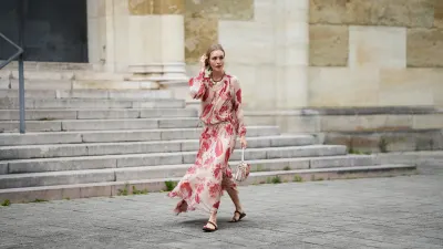 MUNICH, GERMANY - MAY 29: Marlies Pia Pfeifhofer is seen wearing a light beige silk chiffon long sleeve maxi dress with red and pink prints of feathers and flowers, drawstring waist, deep V-neck with front tie and smocking details on the collar and cuffs from FENDI, a natural-colored Baguette canvas bag with coral bandana embroidery, small fringes on the flap, calfskin leather straps, golden hardware and logo detail as well as an additional golden chain metal charm with marine-inspired pendants and a red, pink and white bandana charm with golden logo clip from FENDI, a pair of chocolate brown leather gladiator sandals with a braided leather toe strap and one main strap from Alex Rivière x Manebi, a gold-colored metal and brown nappa leather Forever Fendi necklace with shell-shaped pendants from FENDI, a yellow golden Leaves ring from Ole Lynggaard on May 29, 2025 in Munich, Germany. (Photo by Moritz Scholz/Getty Images)