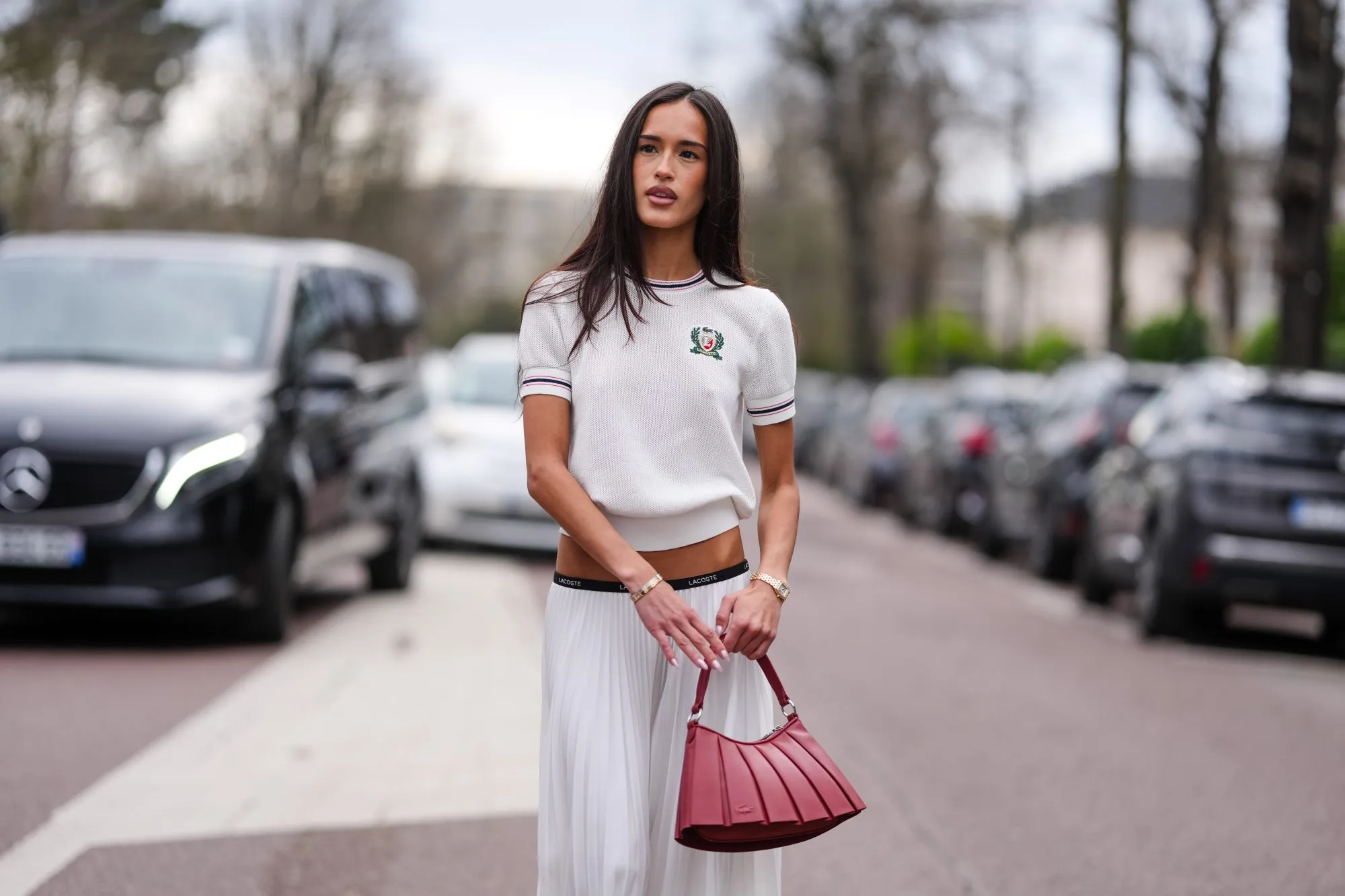 PARIS, FRANCE - MARCH 09: Amalie Bladt wears short sleeve Lacoste sweater, white pleated Lacoste midi skirt, a gold bracelet, a gold watch, shiny dark red Lacoste leather bag, outside Lacoste, during the Paris Fashion week Women's Fall/Winter 2025-2026 on March 9, 2025 in Paris, France. (Photo by Edward Berthelot/Getty Images)
