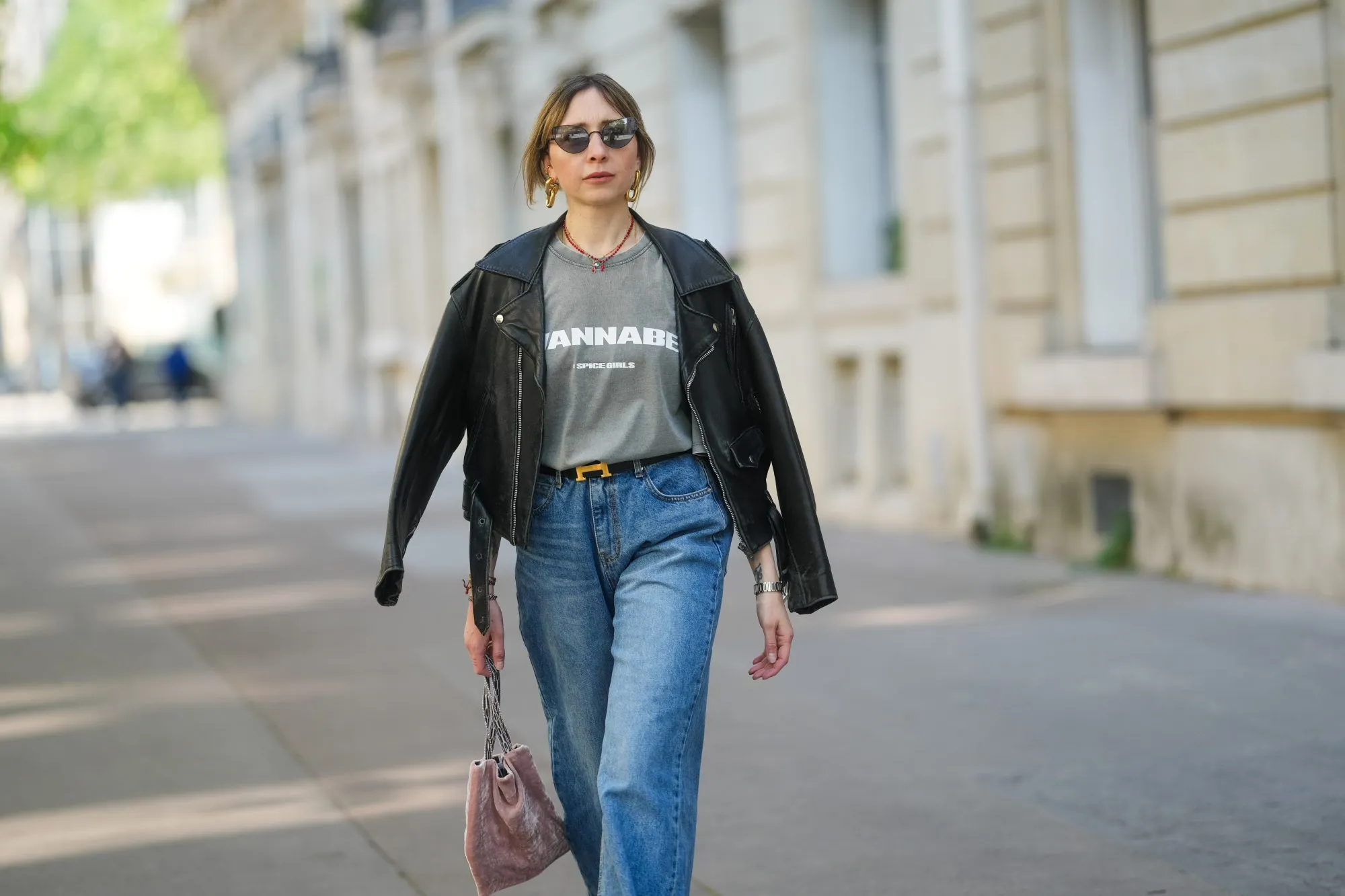 PARIS, FRANCE - APRIL 09: Emy Venturini wears a black leather jacket with wide lapels and zipped pockets, layered over a faded grey graphic T-shirt by Zara featuring &ldquo;WANNABE&rdquo; and the Spice Girls logo. Blue high-waisted jeans by The Frankie Shop and a black belt with a gold H buckle by Hermes are visible. Gold sculptural earrings by Rabanne and a red beaded necklace complete the look. Mid shot, during a street style fashion photo session, on April 09, 2025 in Paris, France. (Photo by Edward Berthelot/Getty Images)
