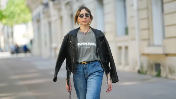 PARIS, FRANCE - APRIL 09: Emy Venturini wears a black leather jacket with wide lapels and zipped pockets, layered over a faded grey graphic T-shirt by Zara featuring “WANNABE” and the Spice Girls logo. Blue high-waisted jeans by The Frankie Shop and a black belt with a gold H buckle by Hermes are visible. Gold sculptural earrings by Rabanne and a red beaded necklace complete the look. Mid shot, during a street style fashion photo session, on April 09, 2025 in Paris, France. (Photo by Edward Berthelot/Getty Images)