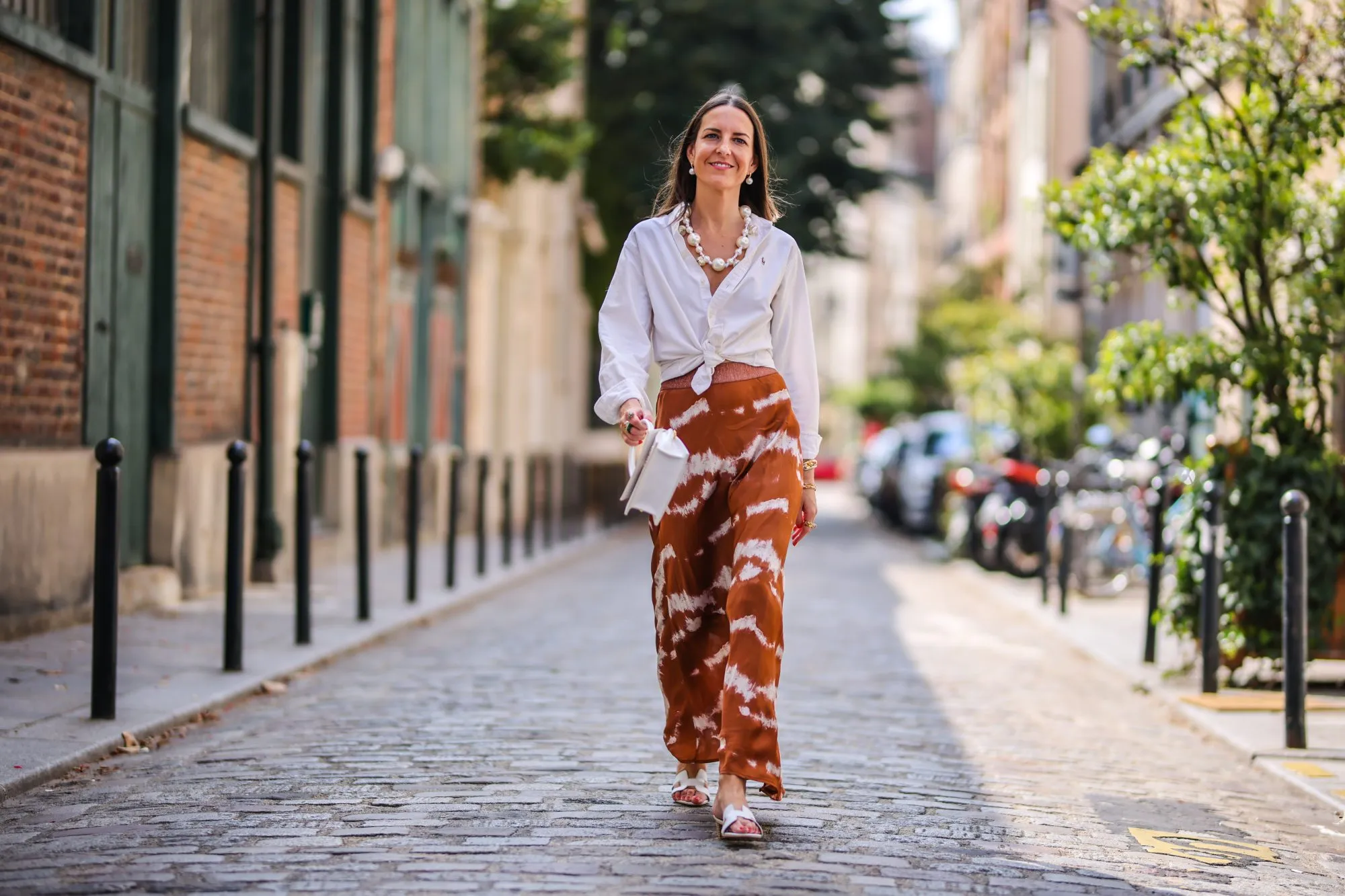 PARIS, FRANCE - JULY 17: Alba Garavito Torre wears a necklace with white large pearls, a white shirt closed with a knot from Ralph Lauren, a brown and white striped tie and dye printed lustrous silky skirt from Imprevu Belgium, white Hermes Oasis sandals, a white Jacquemus bag, on July 17, 2021 in Paris, France. (Photo by Edward Berthelot/Getty Images)