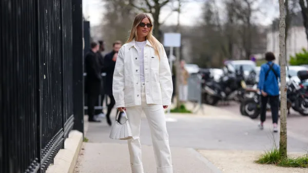 PARIS, FRANCE - MARCH 09: A guest wears dark brown sunglasses, white t-shirt, gold layered necklaces, cream oversized denim jean Lacoste jacket, cream loose denim jean cuffed Lacoste pants, shiny white Lacoste leather bag, dark green heeled leather shoes, outside Lacoste, during the Paris Fashion week Women's Fall/Winter 2025-2026 on March 9, 2025 in Paris, France. (Photo by Edward Berthelot/Getty Images)
