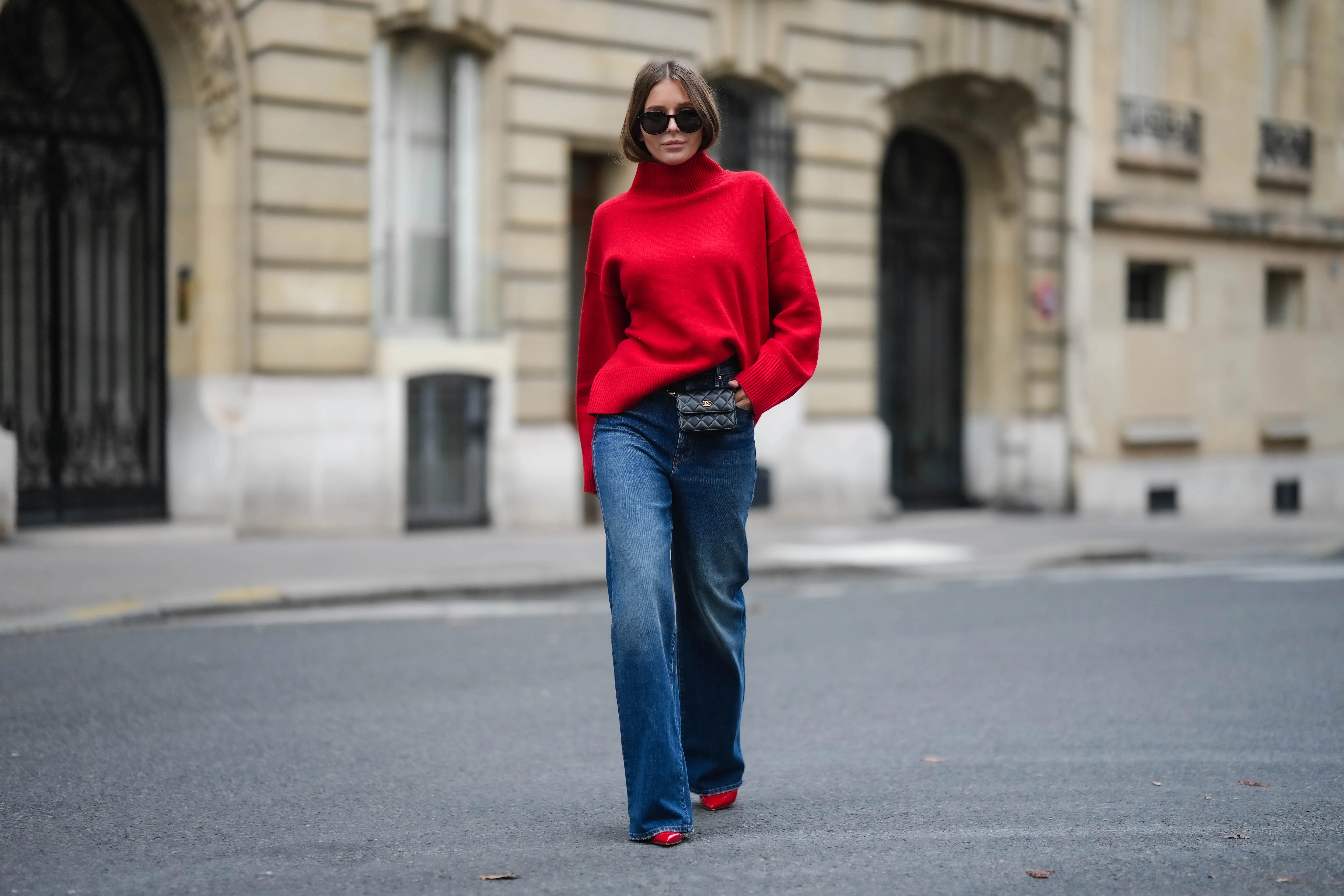 Diane Batoukina wears black sunglasses, a red wool ribbed turtleneck pullover, a black shiny leather belt bag from Chanel, blue faded denim wide legs pants, red shiny varnished leather pointed pumps heels shoes, during a street style fashion photo session, on November 10, 2022 in Paris, France.