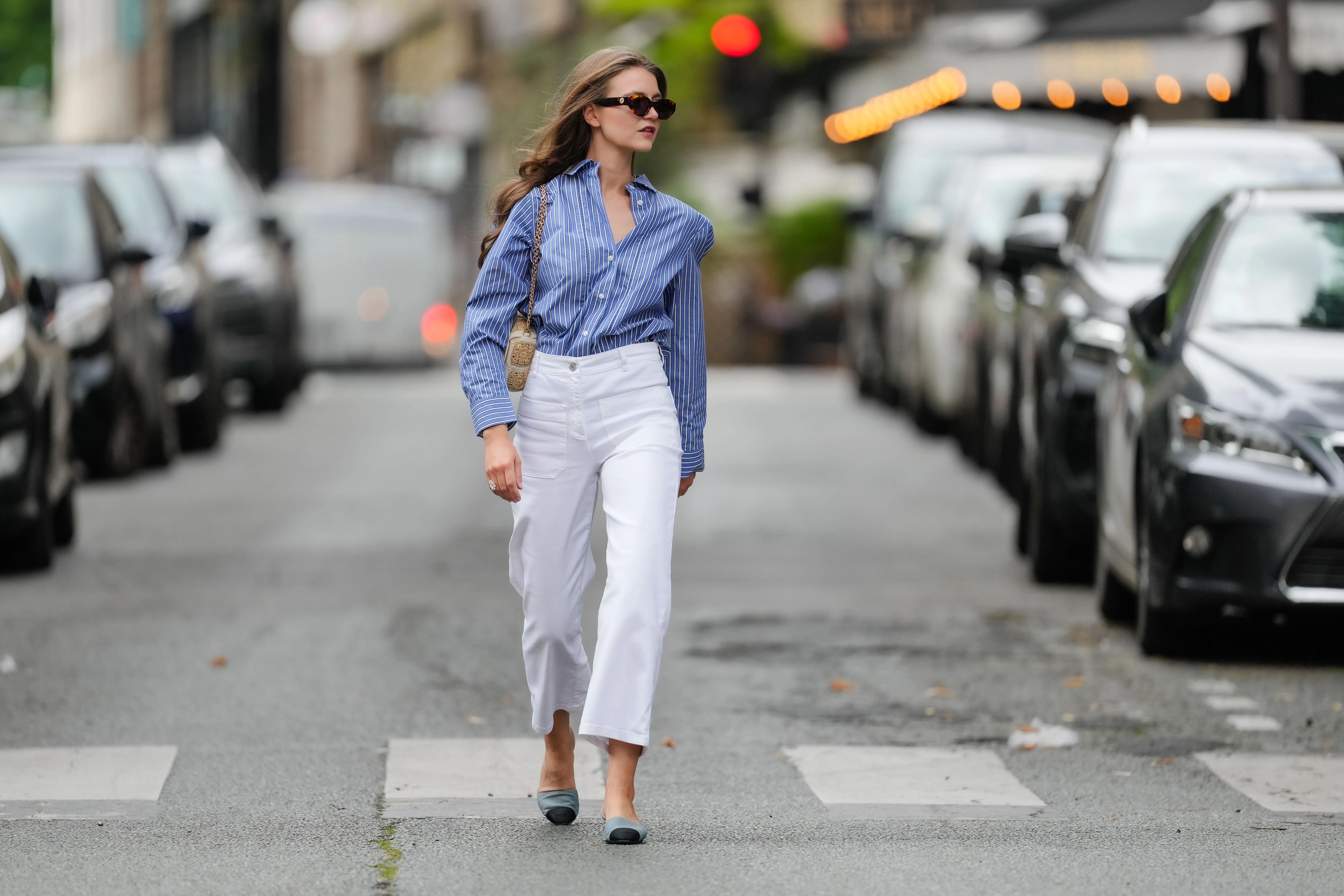 Segolene Hyppolite wears sunglasses, a blue and white oversized shirt from Tommy Hilfiger, a raffia beige bag from Vanessa Bruno, high waist white denim pants from Vanessa Bruno, Chanel slingback shoes in blue denim with black tips, during a street style fashion photo session, on May 20, 2024 in Paris, France.