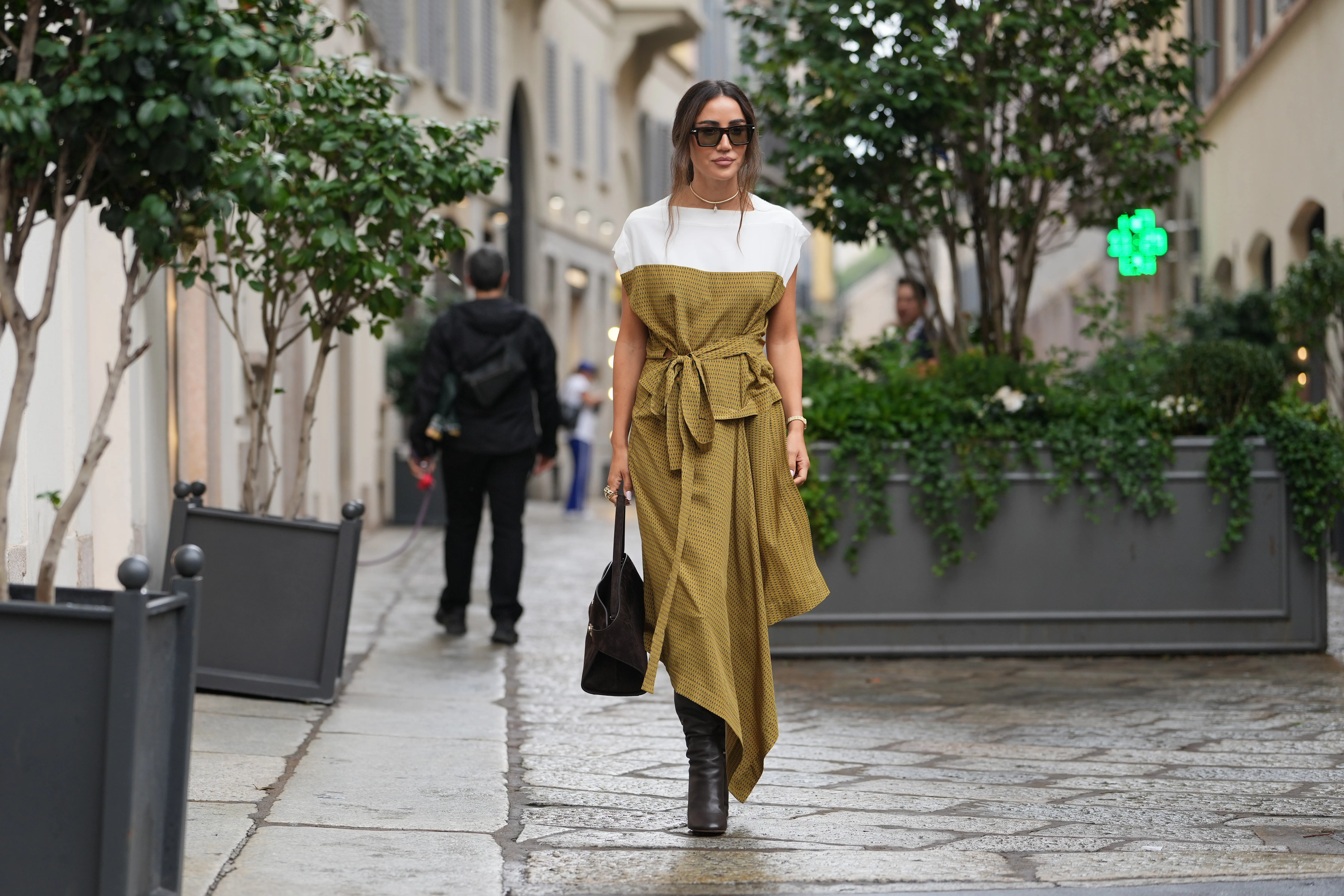 MILAN, ITALY - SEPTEMBER 27: Tamara Kalinic wears long dark brown hair styled in a low loose ponytail with a center part, black rectangular sunglasses, a gold choker-length necklace with matching gold bracelet and rings, a brown suede top-handle satchel bag, a mustard yellow patterned midi dress with a white shoulder yoke and capped sleeves, a self-tie waist belt and an asymmetrical handkerchief hem in a lightweight woven fabric, dark brown leather knee-high boots with a stacked high heel, outside Ferragamo, during Milan Fashion Week - Womenswear Spring/Summer 2026, on September 27, 2025 in Milan, Italy (Photo by Edward Berthelot/Getty Images)