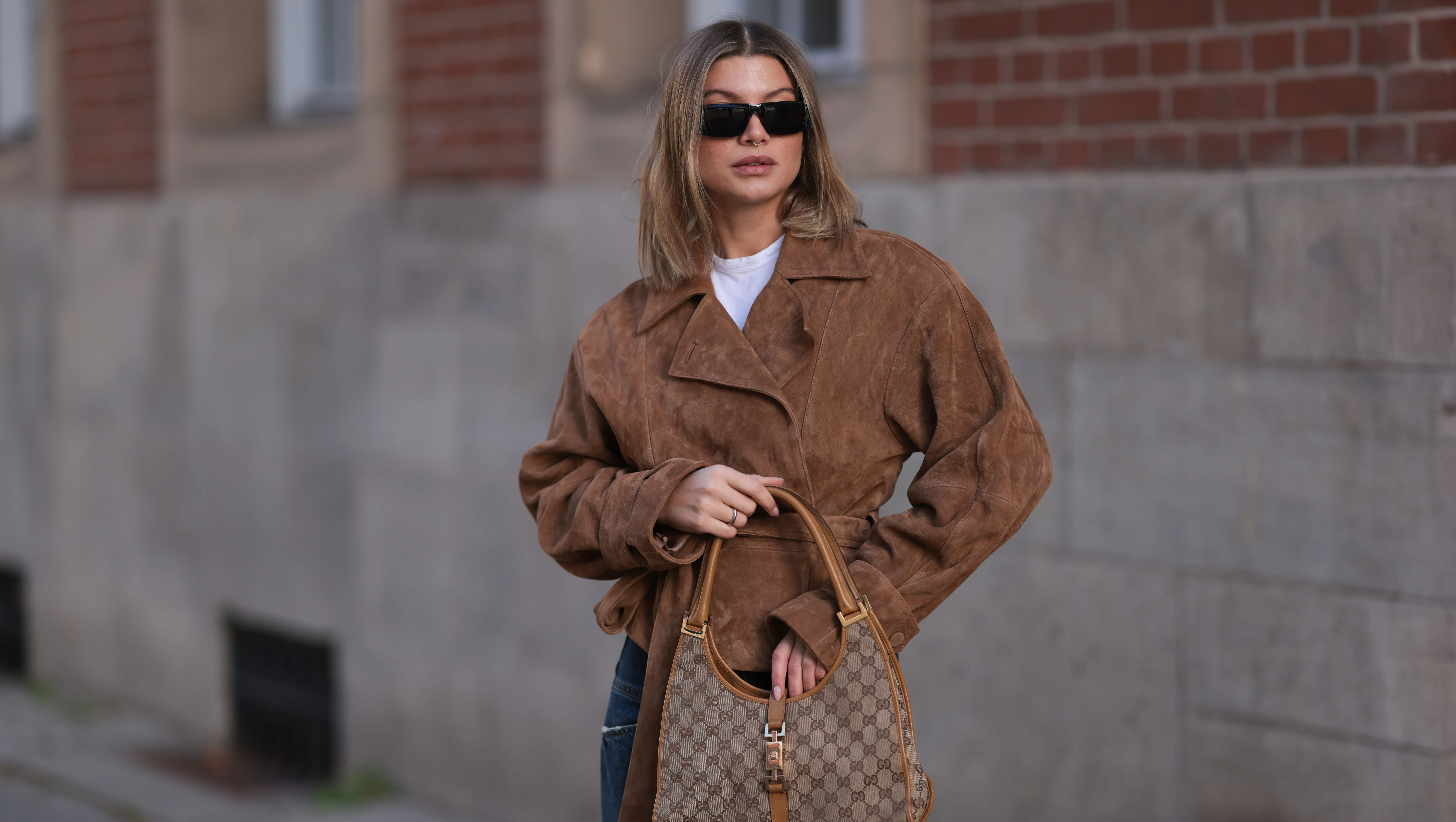 BERLIN, GERMANY - NOVEMBER 02: Sophia Geiss is seen wearing rectangular black sunglasses with a wide tapered temple from Prada; two different-sized gold hoop earrings and a gold stud earring; a silver nose ring; a brown suede jacket with large notch lapels, shoulder pads, decorative seams, a tie belt, and belted cuffs from & Other Stories; a white t-shirt underneath; a brown logo-embellished canvas bag with brown leather details and handles from Gucci; a silver ring; ans wide-legged dark jeans with orange decorative seams from Closed on November 02, 2024 in Berlin, Germany. (Photo by Jeremy Moeller/Getty Images)