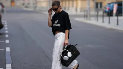 BERLIN, GERMANY - JULY 27: Sophia Geiss is seen wearing a black oversized t-shirt with bold white logo print across the chest worn tucked in her bra to create a cropped look from Eme Studios; white cotton parachute pants with a wide-leg silhouette and gathered elastic cuffs from Arket; a black leather The Hudson bag with gold-tone hardware detail, belted detail and top handles from Demellier London; a pair of white Apple AirPods Max are casually adorned to the bags top handles;black Havaianas Slim rubber flip-flops with a classic thong design from Havaianas; big black oval sunglasses with a glossy acetate frame from Mango; a small golden bangle bracelet from Daniel Wellington; her brown hair is worn in a sleek low bun on July 27, 2025 in Berlin, Germany. (Photo by Moritz Scholz/Getty Images)