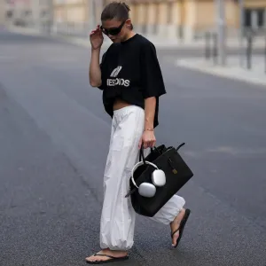 BERLIN, GERMANY - JULY 27: Sophia Geiss is seen wearing a black oversized t-shirt with bold white logo print across the chest worn tucked in her bra to create a cropped look from Eme Studios; white cotton parachute pants with a wide-leg silhouette and gathered elastic cuffs from Arket; a black leather The Hudson bag with gold-tone hardware detail, belted detail and top handles from Demellier London; a pair of white Apple AirPods Max are casually adorned to the bags top handles;black Havaianas Slim rubber flip-flops with a classic thong design from Havaianas; big black oval sunglasses with a glossy acetate frame from Mango; a small golden bangle bracelet from Daniel Wellington; her brown hair is worn in a sleek low bun on July 27, 2025 in Berlin, Germany. (Photo by Moritz Scholz/Getty Images)