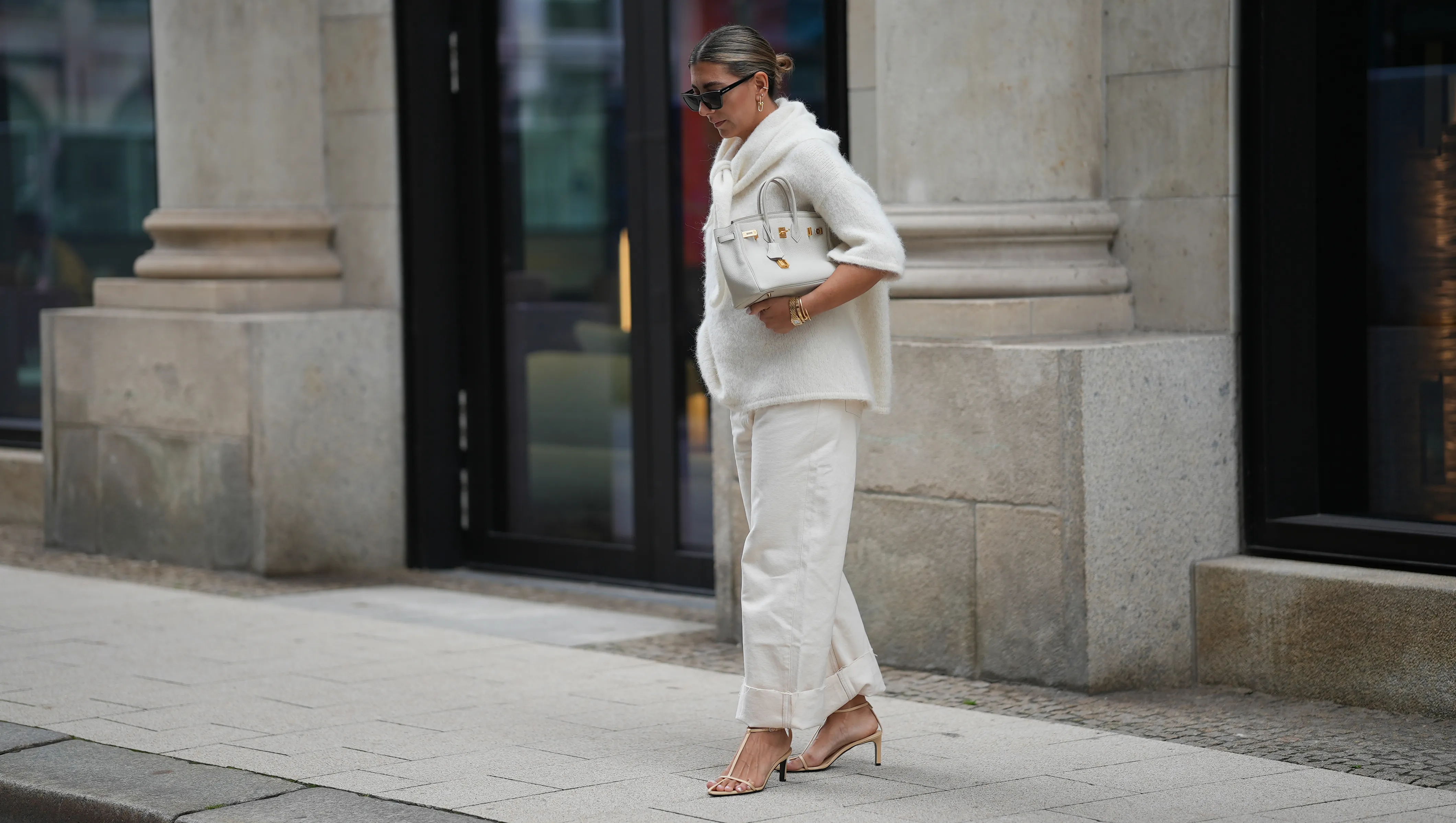 HAMBURG, GERMANY - SEPTEMBER 19: Aylin K&radic;&part;nig is seen wearing a short-sleeved, fluffy textured, oversized sweater in off-white with a high, tied neckline; relaxed off-white jeans with a wide cuff at the hem; nude strappy heeled sandals; a medium-sized white leather handbag with gold hardware; black rectangular sunglasses; gold link earrings from Tiffany & Co HardWear Collection; detailed gold bracelets from Cartier; a watch with a metallic wristband and white clock face from Cartier; her dark hair is pulled back into a neat, low bun on September 19, 2025 in Hamburg, Germany. (Photo by Moritz Scholz/Getty Images)