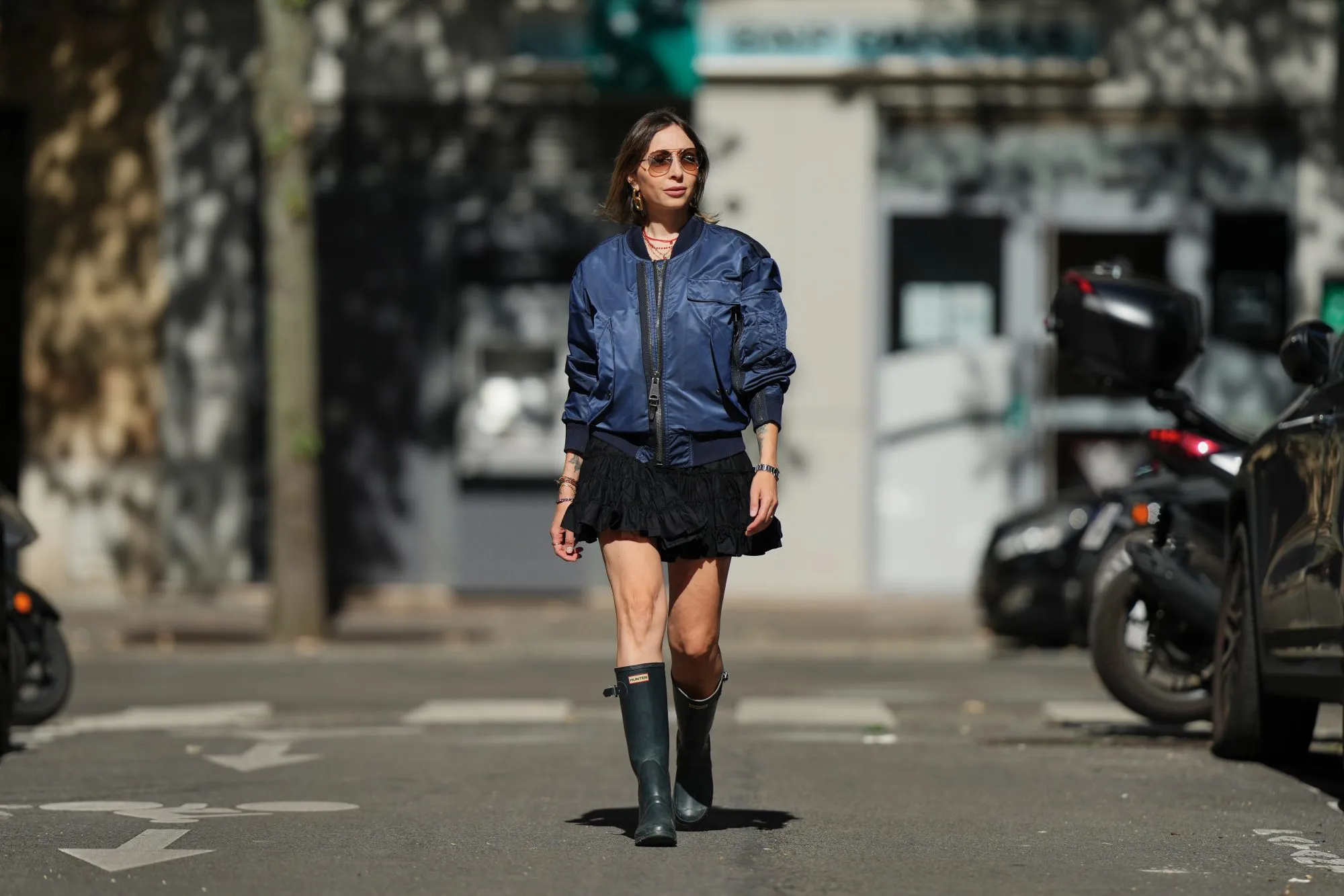 PARIS, FRANCE - SEPTEMBER 06: Emy Venturini wears straight light brown hair with a middle part, brown gradient sunglasses by Prada, gold hoop earrings by Rabanne, layered necklaces, multiple bracelets, a navy blue bomber jacket by Burberry, a short ruffled black dress by Zara Studio, and dark green rubber rain boots by Hunter, during a street style fashion photo session, on September 06, 2025 in Paris, France. (Photo by Edward Berthelot/Getty Images)