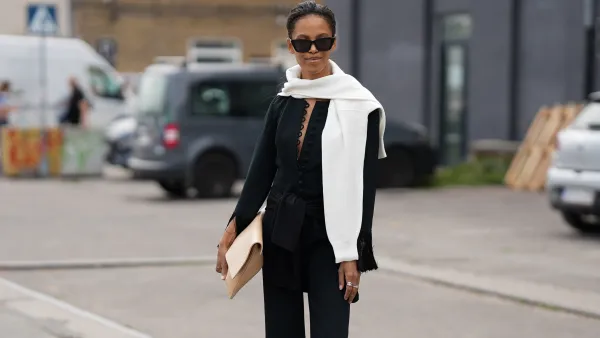 COPENHAGEN, DENMARK - AUGUST 7: Suzannà Gembege is seen wearing a white knit sweater draped over the shoulders; a black blouse with a deep V-neck big button details and fringe-accented sleeves; a matching black fringe-hem trousers; black flip flops; a black cat-eye sunglasses; a beige oversized clutch; silver rings; her black hair is styled straight in a ponytail outside the Marimekko show during Copenhagen Fashion Week day four on August 7, 2025 in Copenhagen, Denmark. (Photo by Moritz Scholz/Getty Images)