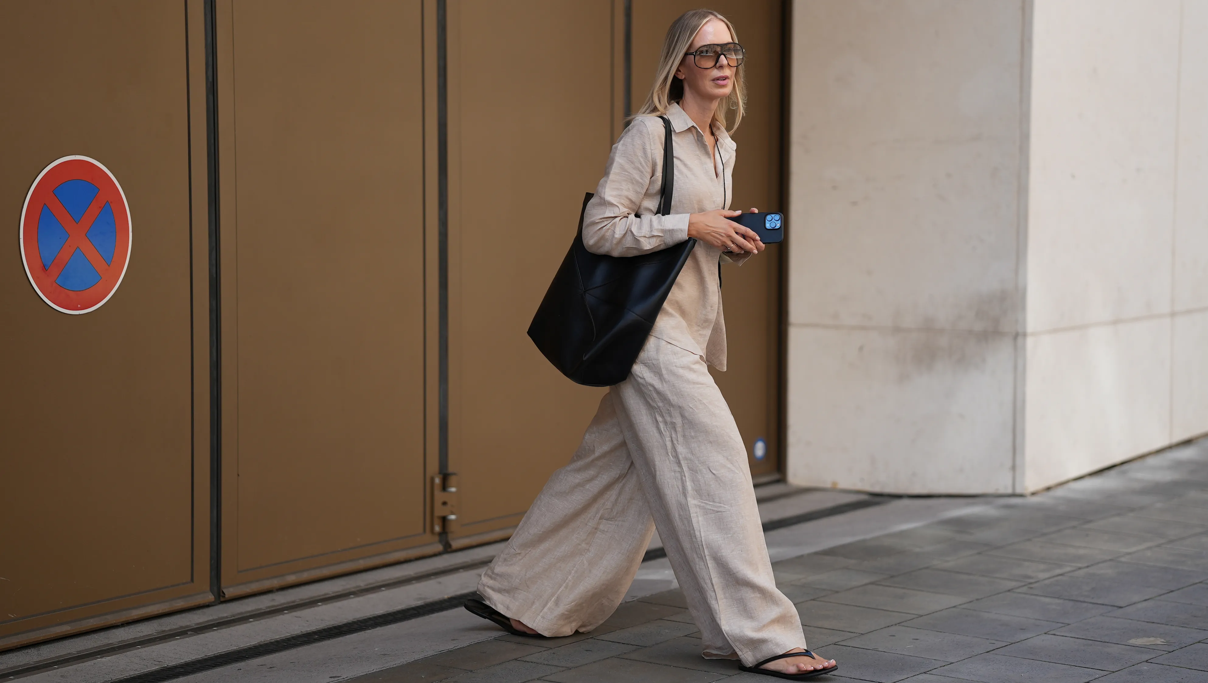 MUNICH, GERMANY - AUGUST 8: Mirja Klein is seen wearing a relaxed beige linen shirt with a pointed collar, hidden button-down front and long sleeves from Falconeri; wide-leg beige linen trousers with pleats from Falconeri; a pair of minimalistic rubber flat flip flop sandals from A.Emery; a large black leather medium Puzzle Fold Tote bag with geometric stitching, double shoulder straps and open-top design from Loewe; oversized square mask sunglasses with a tortoiseshell acetate frame and gradient lenses from Tom Ford on August 8, 2025 in Munich, Germany. (Photo by Moritz Scholz/Getty Images)