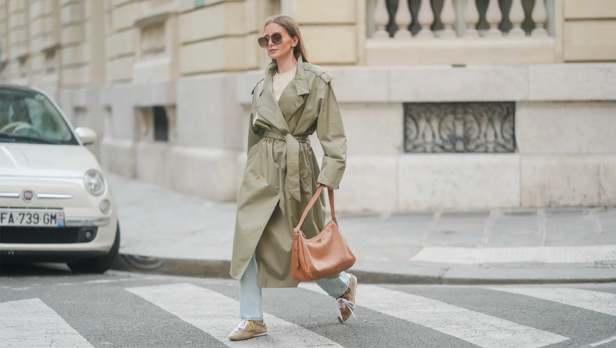 Diane Batoukina wears her long straight brown hair down with a center part, oversized square Dior sunglasses, a khaki belted trench coat by Sandro layered over a beige T-shirt by Zara, light-wash straight-leg jeans by Anine Bing, beige suede Miu Miu sneakers with mismatched laces and tassel details, and carries a large tan leather shoulder bag by Prada, walking across a pedestrian crossing, during a street style fashion photo session on March 25, 2025 in Paris, France.
