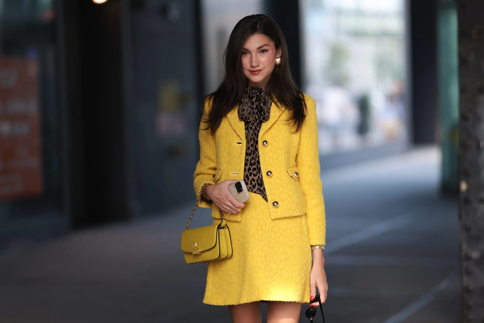 NEW YORK, NEW YORK - SEPTEMBER 08: A Fashion Week guest is seen wearing black sunglasses, golden earrings, a yellow matching two-piece from Kate Spade consisting of a jacket with golden buttons and a mini skirt, underneath a brown/black top with animal print and a bow, a silver/golden watch, a yellow leather bag from Kate Spade and pumps with ankle strap and animal print from Kate Spade outside before Kate Spade presentation on September 08, 2023 in New York City. (Photo by Jeremy Moeller/Getty Images)