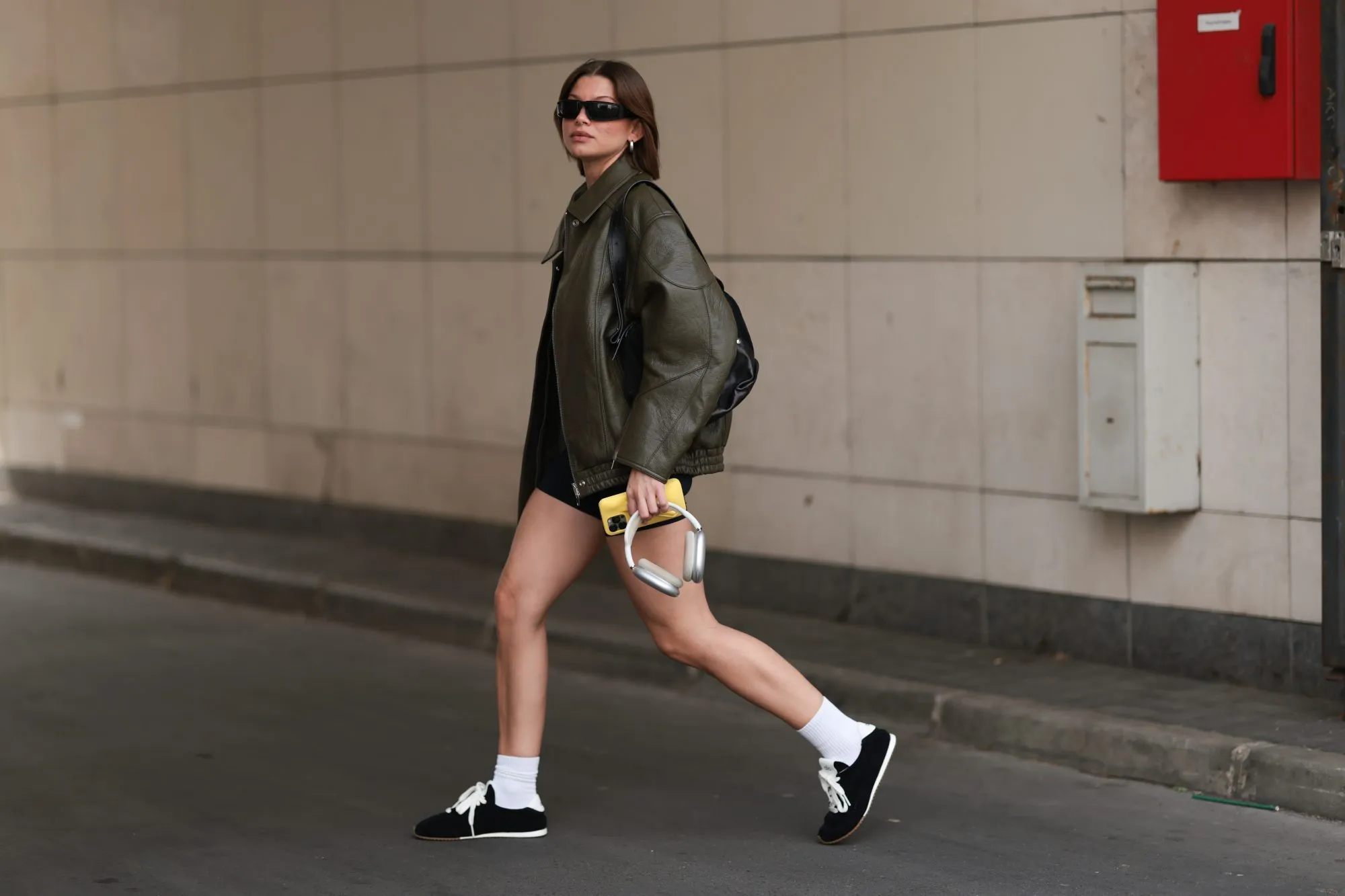 BERLIN, GERMANY - AUGUST 10: Sophia Geiss is seen wearing rectangular black sunglasses with wide tapered temples from Prada, chunky silver hoop earrings, a silver nose ring, an oversized olive faux leather jacket with drop shoulders and a pointed collar from The Frankie Shop, a fitted black vintage romper with short legs, a black leather shoulder bag, white socks, and black suede sneakers with brown rubber soles from Alo, while holding silver Apple AirPods Max headphones and an Apple iPhone with a yellow lip iPhone case from Rhode on August 10, 2025 in Berlin, Germany. (Photo by Jeremy Moeller/Getty Images)