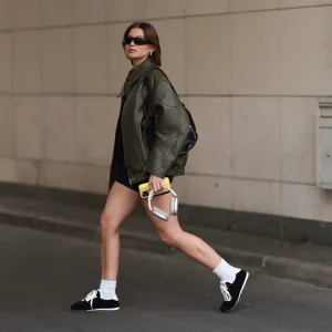BERLIN, GERMANY - AUGUST 10: Sophia Geiss is seen wearing rectangular black sunglasses with wide tapered temples from Prada, chunky silver hoop earrings, a silver nose ring, an oversized olive faux leather jacket with drop shoulders and a pointed collar from The Frankie Shop, a fitted black vintage romper with short legs, a black leather shoulder bag, white socks, and black suede sneakers with brown rubber soles from Alo, while holding silver Apple AirPods Max headphones and an Apple iPhone with a yellow lip iPhone case from Rhode on August 10, 2025 in Berlin, Germany. (Photo by Jeremy Moeller/Getty Images)