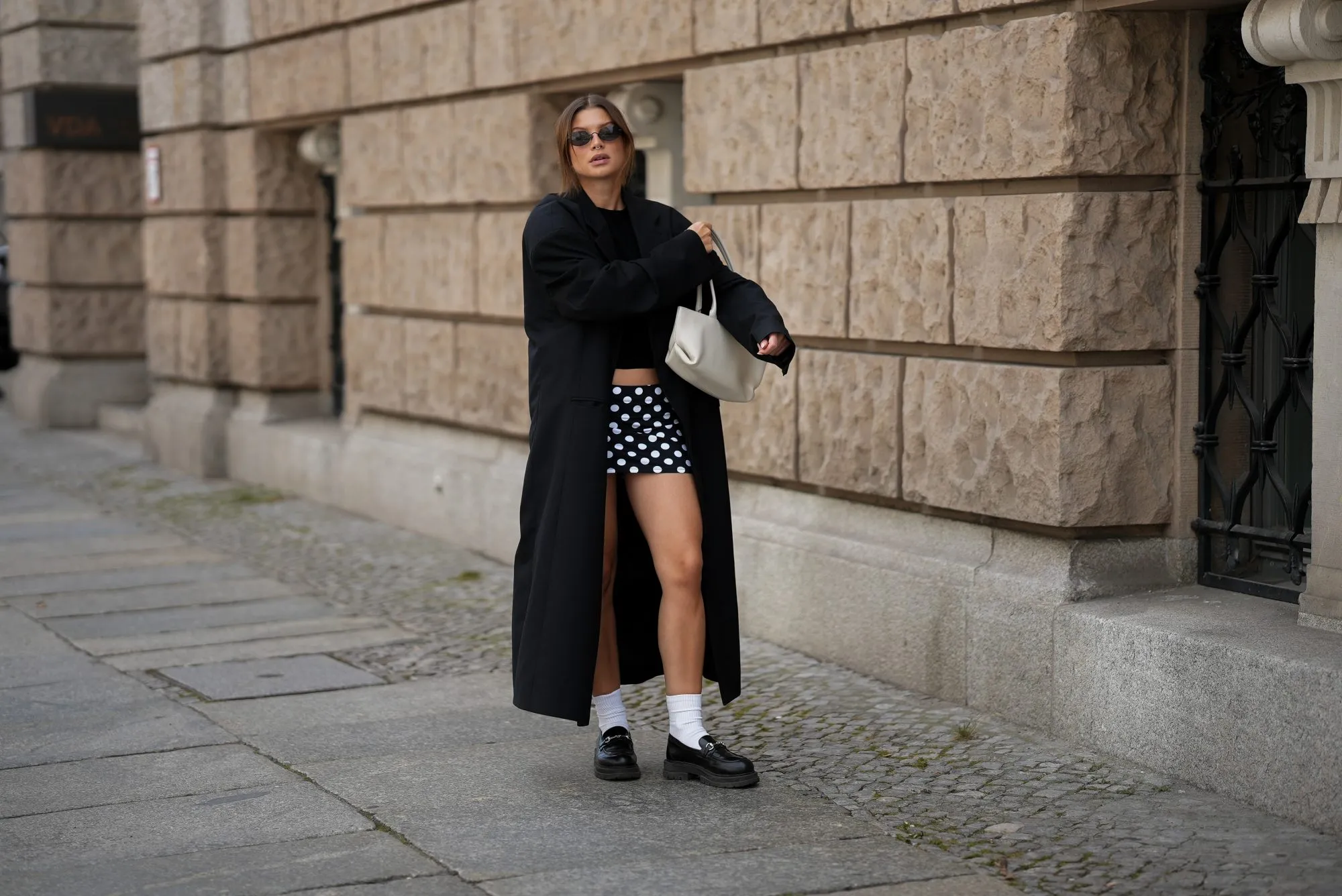 BERLIN, GERMANY - SEPTEMBER 1: Sonia Lyson is seen wearing a long, oversized black coat with broad shoulders and a straight cut, worn open from 032C; a simple black crop top leaving the midriff slightly visible from Zara; a short, fitted black-and-white polka dot mini skirt from Attega; black chunky loafers paired with white socks from LeGer by Lena Gercke; a medium sized white leather handbag with shoulder straps from Demmilie London; small, sleek black sunglasses that are oval shaped from Vela Eyewear; her brown hair is pulled back into a slightly messy low bun on September 1, 2025 in Berlin, Germany. (Photo by Moritz Scholz/Getty Images)