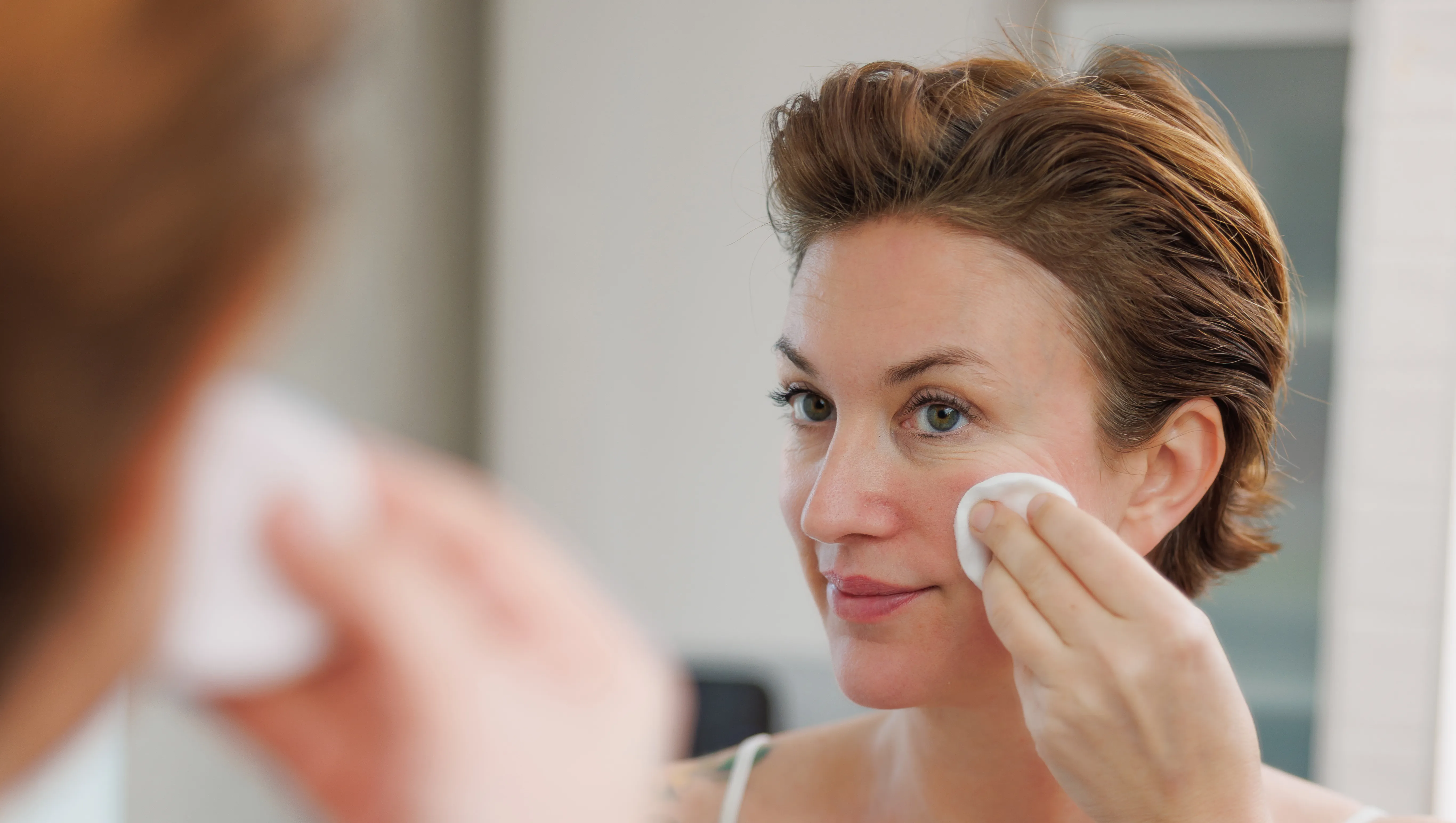 Beautiful mid adult woman with short hair wearing camisole doing her skin care routine in the morning, applying facial cleanser with pad in front of the bathroom mirror