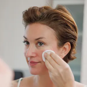 Beautiful mid adult woman with short hair wearing camisole doing her skin care routine in the morning, applying facial cleanser with pad in front of the bathroom mirror