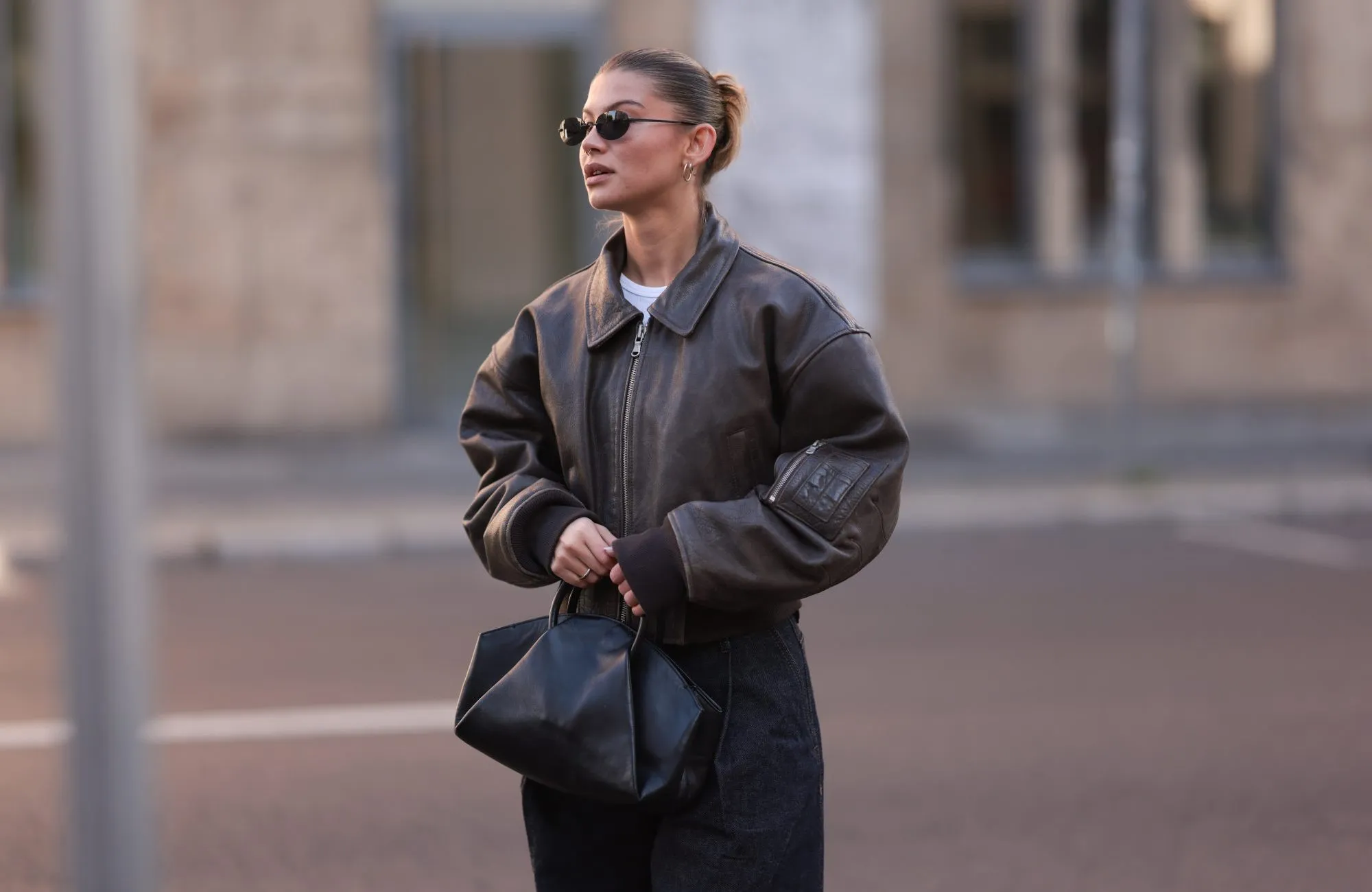 BERLIN, GERMANY - OCTOBER 12: Sophia Geiss is seen wearing oval rimless black sunglasses from Vehla Eyewear, two different-sized gold hoop earrings, a silver nose ring, a cropped dark brown leather jacket with wide sleeves and ribbed cotton cuffs from Meotine, a white top underneath, wide-legged dark vintage jeans with orange decorative seams, a silver ring, and a black leather bag with round handles from COS on October 12, 2024 in Berlin, Germany. (Photo by Jeremy Moeller/Getty Images)
