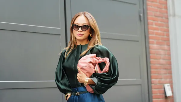 MILAN, ITALY - SEPTEMBER 24: A guest wears long straight light brown hair with a center part, black cat-eye sunglasses, gold hoop earrings and gold bracelets, a pink leather shoulder bag with a knotted handle, a dark green satin blouse with a high neckline and voluminous long puff sleeves, high-waisted deep teal satin wide-leg trousers secured with a black leather belt from Fendi with a gold buckle, outside Fendi, during Milan Fashion Week - WoWomenswear Spring/Summer 2026, on September 24, 2025 in Milan, Italy (Photo by Edward Berthelot/Getty Images)