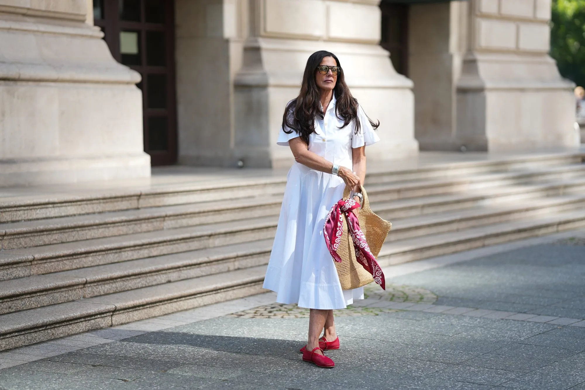 FRANKFURT AM MAIN, GERMANY - JULY 10: Angelika Hanel is seen wearing a white short-sleeved shirt midi dress with a flared silhouette and soft pleating from Erika Cavallini; a pair of signature Alaïa ballet flats in red fishnet textile and patent leather, a buckle strap on top and round toes from Alaïa; the ICARE maxi shopper crafted from hand-embroidered natural raffia and a brown gold oversized, sculptured Cassandre from Saint Laurent adorned with a red and white silk bandana from Arket tied to the handle; a silver Rolex Daytona watch; a yellow golden Juste un Clou bracelet from Cartier; a pavé diamond Tennis bracelet; a bold sterling silver Bone Cuff from Tiffany & Co.; oversized black aviator sunglasses with orange shaded glasses from Kapten & Son; long brown hair and natural make-up on July 10, 2025 in Frankfurt am Main, Germany. (Photo by Moritz Scholz/Getty Images)