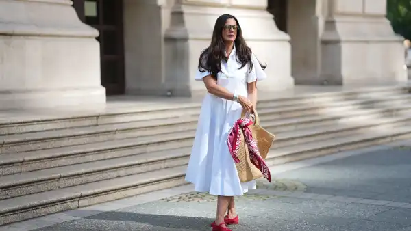 FRANKFURT AM MAIN, GERMANY - JULY 10: Angelika Hanel is seen wearing a white short-sleeved shirt midi dress with a flared silhouette and soft pleating from Erika Cavallini; a pair of signature Alaïa ballet flats in red fishnet textile and patent leather, a buckle strap on top and round toes from Alaïa; the ICARE maxi shopper crafted from hand-embroidered natural raffia and a brown gold oversized, sculptured Cassandre from Saint Laurent adorned with a red and white silk bandana from Arket tied to the handle; a silver Rolex Daytona watch; a yellow golden Juste un Clou bracelet from Cartier; a pavé diamond Tennis bracelet; a bold sterling silver Bone Cuff from Tiffany & Co.; oversized black aviator sunglasses with orange shaded glasses from Kapten & Son; long brown hair and natural make-up on July 10, 2025 in Frankfurt am Main, Germany. (Photo by Moritz Scholz/Getty Images)
