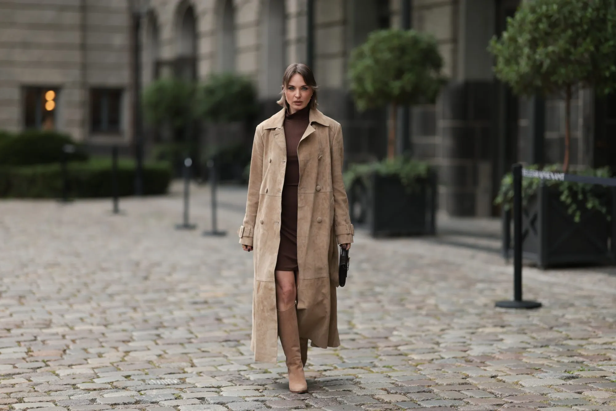 BERLIN, GERMANY - SEPTEMBER 17: Yana Pries seen wearing a long suede leather trench coat from NOUR HAMMOUR in a warm beige tone. The coat features double-breasted buttons, a structured collar, and belted cuffs, creating a luxurious yet timeless silhouette. Her hair is cut into a sleek bob, light brown with subtle highlights, styled with a middle part and softly curved ends that frame her face. Underneath she wears Even&Odd fitted chocolate-brown mini dress, with a high neckline, Bottega Veneta Andiamo clutch, a structured black leather bag with a gold knot detail on the strap and Bobbies knee-high Bobbies suede leather boots in a soft beige tone, on September 17, 2025 in Berlin, Germany. (Photo by Jeremy Moeller/Getty Images)