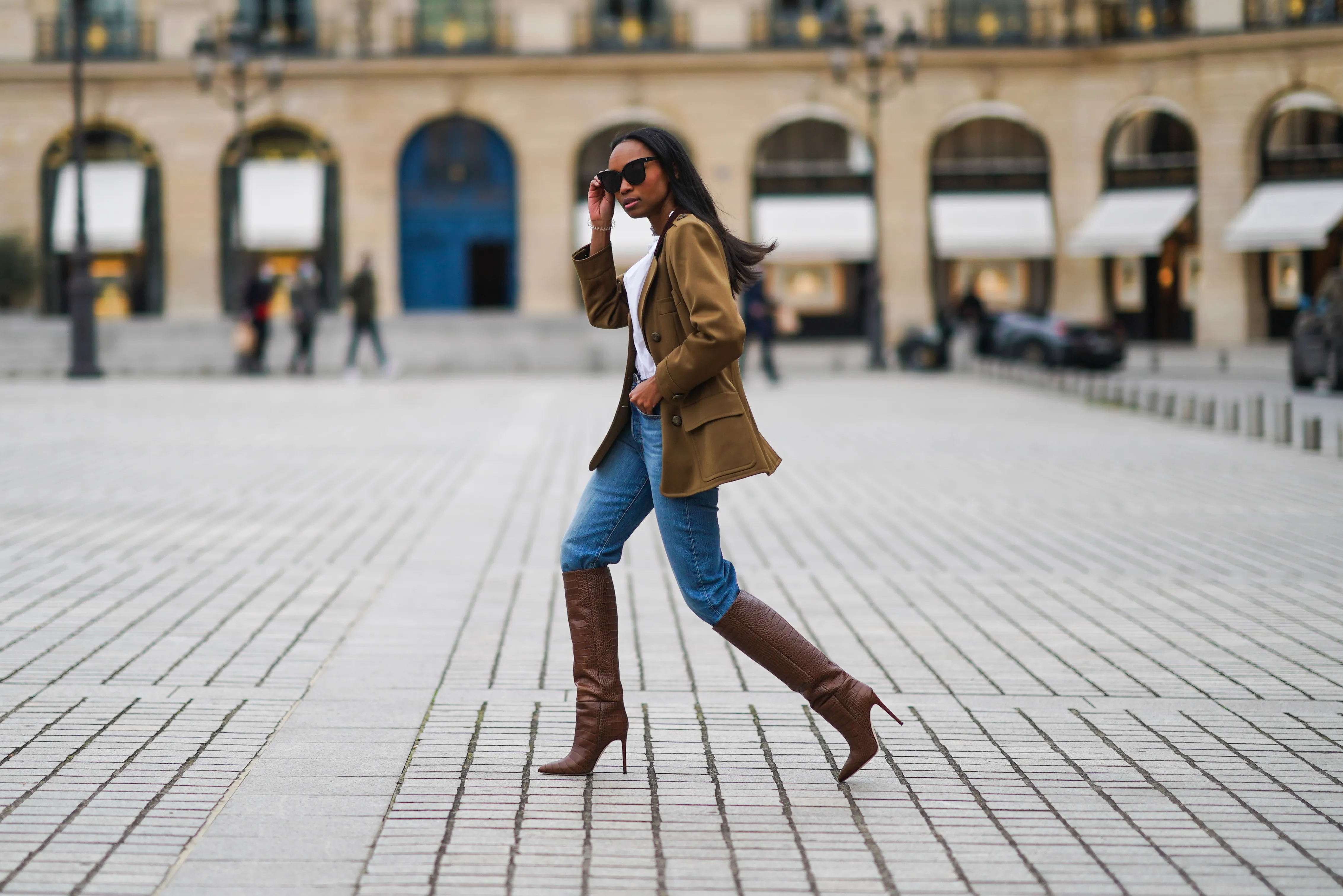 Emilie Joseph wears sunglasses, a khaki military jacket from Saint-Laurent YSL, a white shirt / blouse from Zara with ruffled collar, blue denim jeans pants from Levi's, brown high heeled knee-high pointy leather crocodile pattern boots from Paris Texas, on January 26, 2021 in Paris, France.