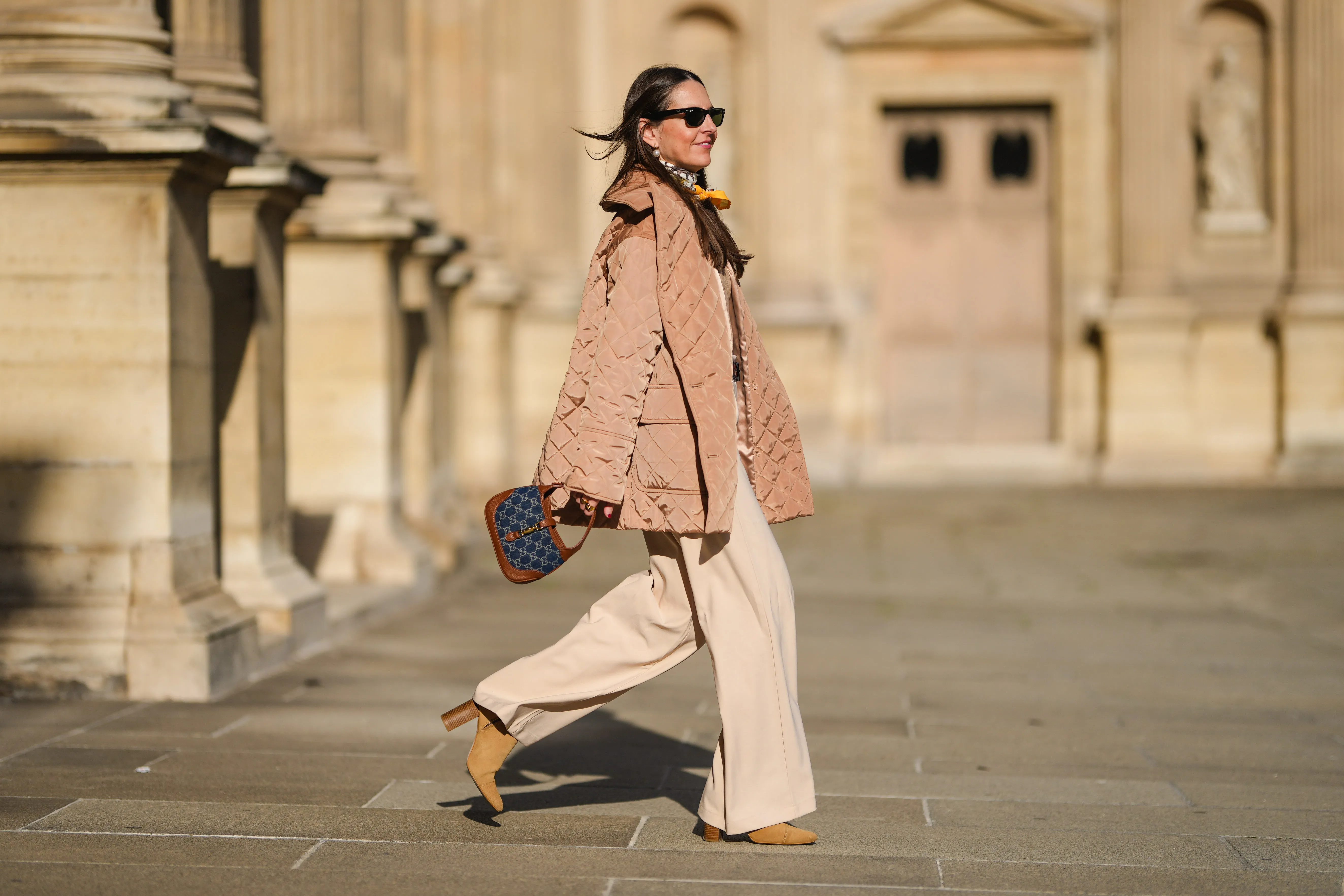 Alba Garavito Torre wears pearls earrings, black wayfarer sunglasses from Ray Ban, a beige quilted oversized jacket from Munthe, a white and yellow silk scarf tie with classic knot from Lollys Laundry, a brown / beige braided wool pullover from Polo Ralph Lauren, a brown shiny leather belt, high waist beige palazzo pants from Bimba y Lola, a blue denim monogram and brown shiny leather Jackie 1961 Mini bag handbag from Gucci, yellow mustard suede block heels ankle boots, gold bracelets, gold rings, during a street style fashion photo session, on November 12, 2021 in Paris, France.