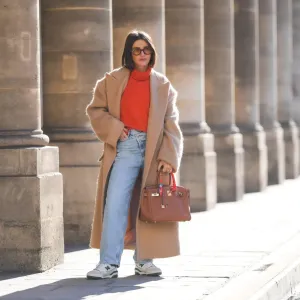 PARIS, FRANCE - FEBRUARY 09: Alexandra Pereira wears brown sunglasses, gold earrings, a neon orange turtleneck wool pullover, a beige oversized long winter coat, a brown shiny leather Birkin handbag from Hermes, diamonds ring, an orange / blue / yellow / white print pattern silk scarf from Hermes, blue faded denim large mom jeans pants, white and green leather sneakers from New Balance, during a street style fashion photo session, on February 09, 2022 in Paris, France. (Photo by Edward Berthelot/Getty Images)
