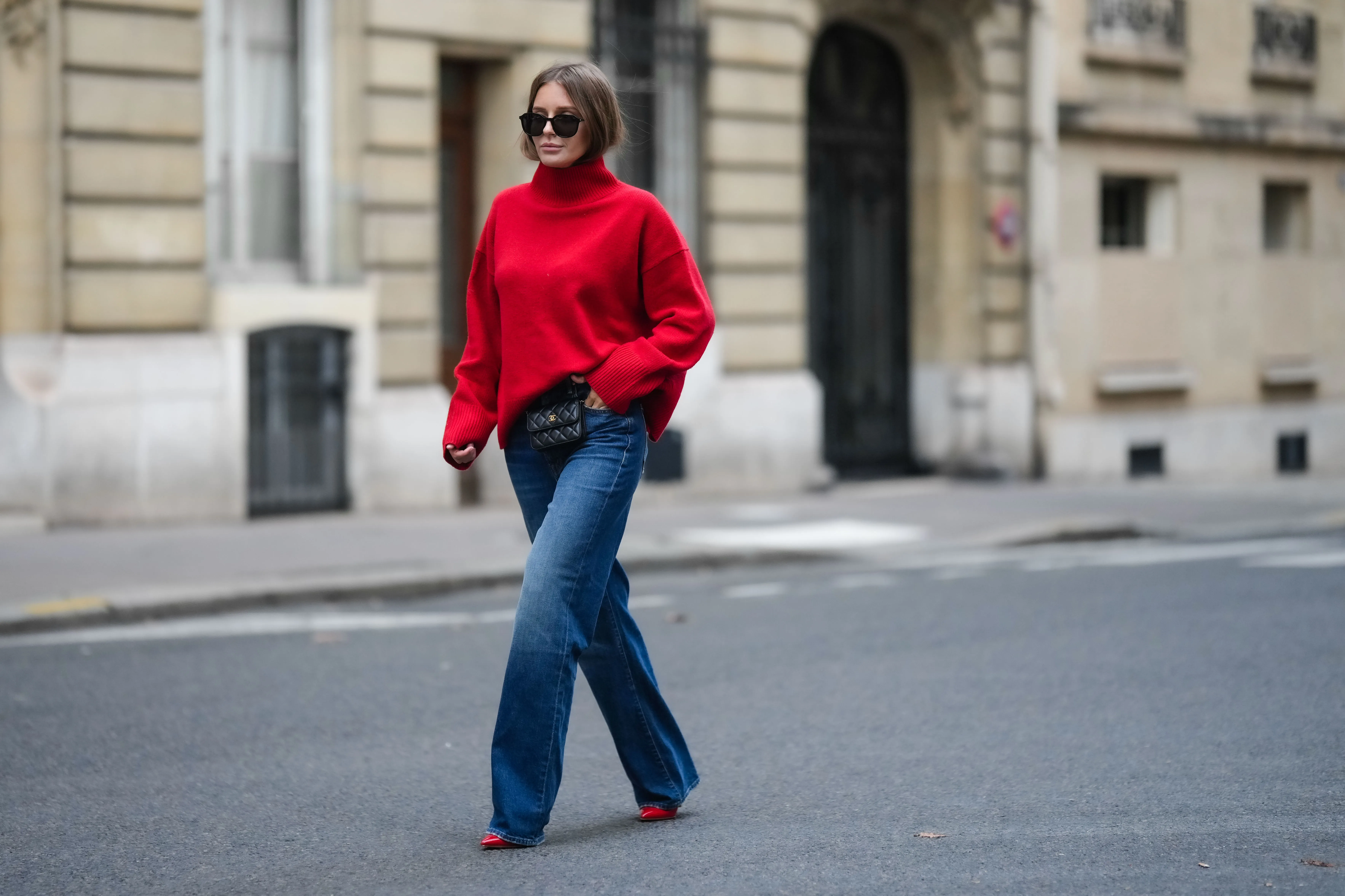 Diane Batoukina wears black sunglasses, a red wool ribbed turtleneck pullover, a black shiny leather belt bag from Chanel, blue faded denim wide legs pants, red shiny varnished leather pointed pumps heels shoes, during a street style fashion photo session, on November 10, 2022 in Paris, France.