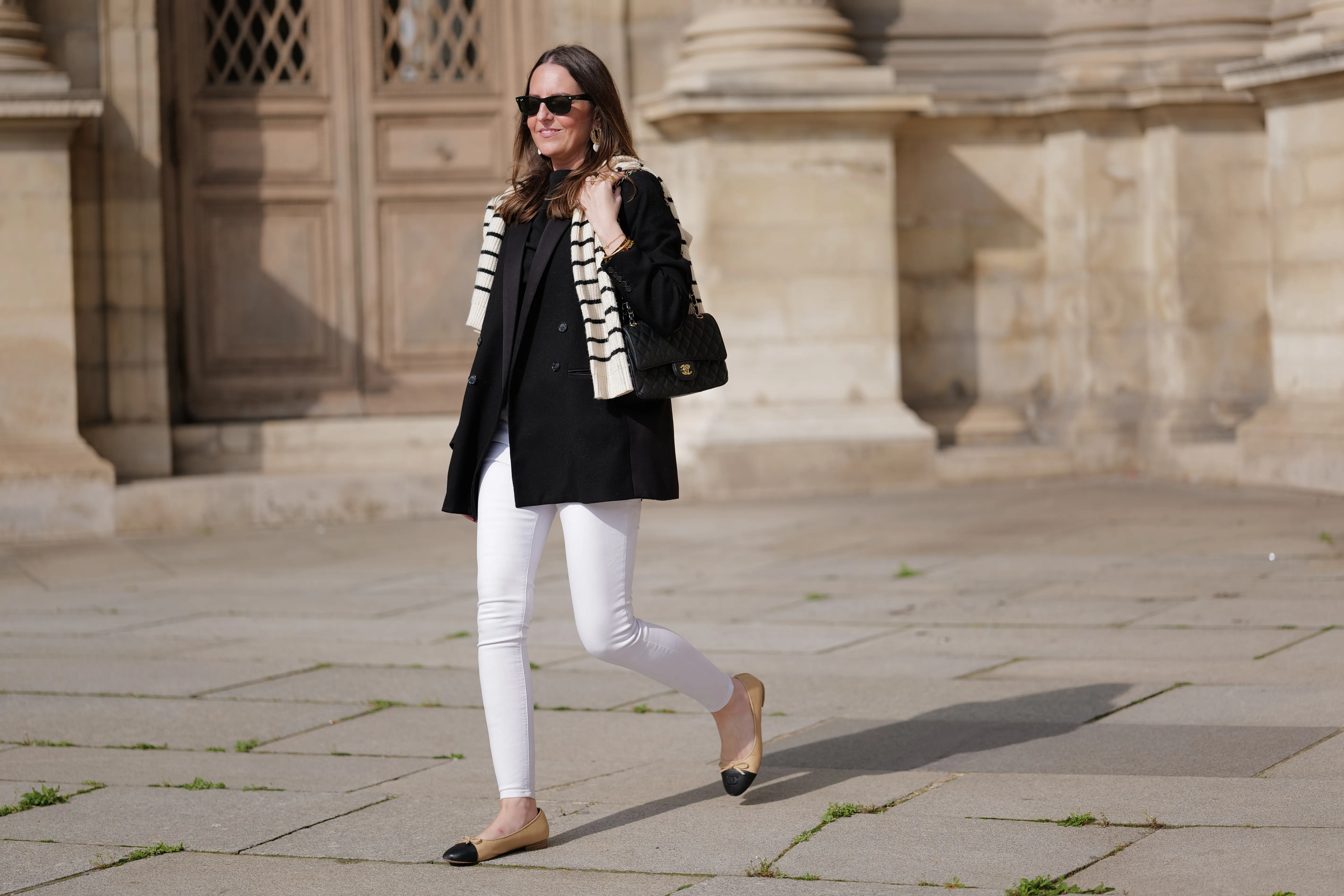 Alba Garavito Torre wears black sunglasses from Ray Ban, gold and white pendant large earrings, a black t-shirt, a white with black small striped print pattern wool pullover from Massimo Dutti, a black shiny grained leather Timeless shoulder bag from Chanel, a black oversized blazer jacket from Munthe, white skinny denim pants from Zara, beige shiny leather with black toe-cap ballerinas from Chanel , during a street style fashion photo session, on April 29, 2023 in Paris, France.