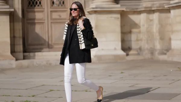 Alba Garavito Torre wears black sunglasses from Ray Ban, gold and white pendant large earrings, a black t-shirt, a white with black small striped print pattern wool pullover from Massimo Dutti, a black shiny grained leather Timeless shoulder bag from Chanel, a black oversized blazer jacket from Munthe, white skinny denim pants from Zara, beige shiny leather with black toe-cap ballerinas from Chanel , during a street style fashion photo session, on April 29, 2023 in Paris, France.