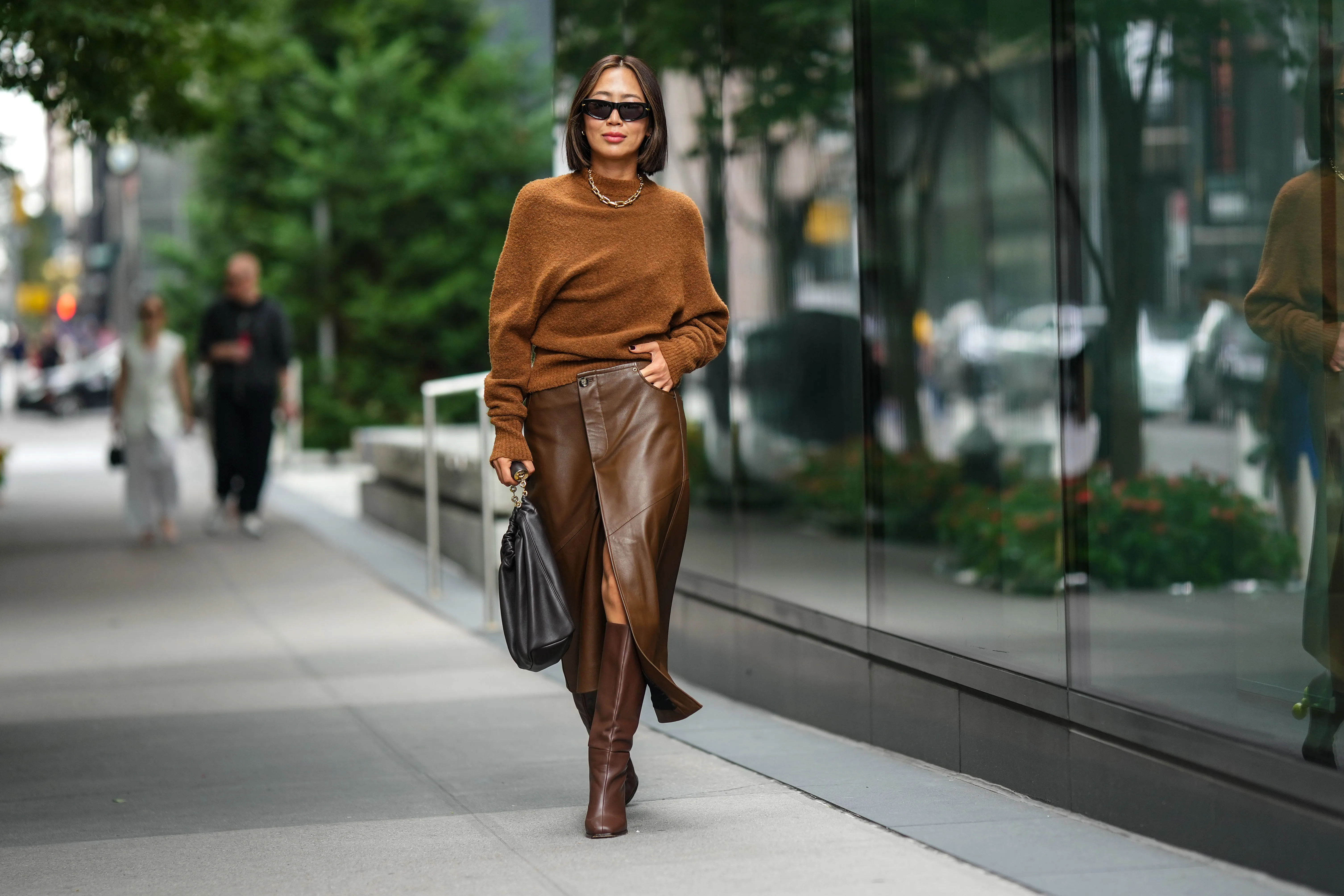Aimee Song wears sunglasses, a necklace, a brown wool pullover, a brown leather skirt, a black leather bag, brown knee-high boots, outside Proenza Schouler, during New York Fashion Week, on September 09, 2023 in New York City.