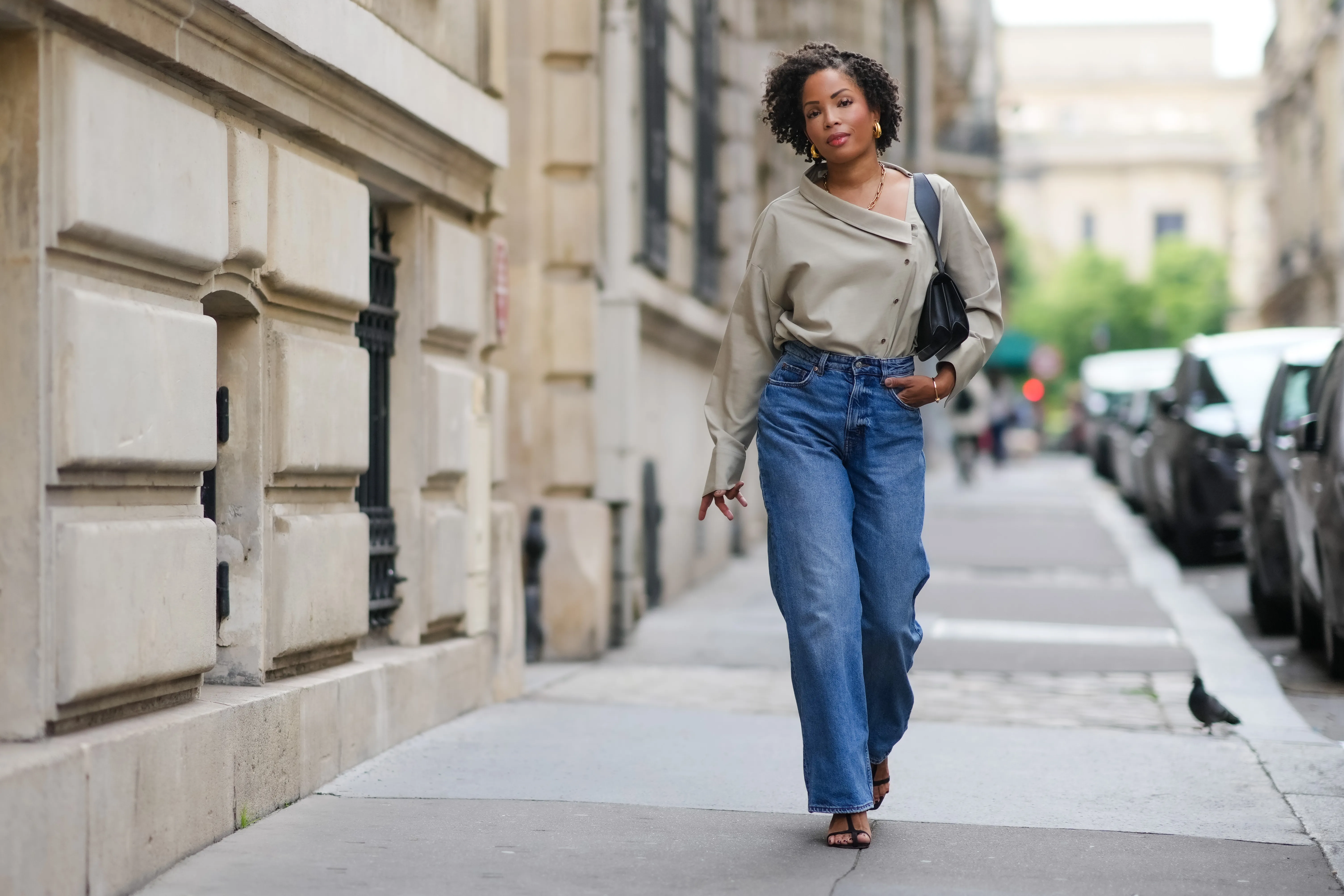 PARIS, FRANCE - MAY 20: Ellie Delphine wears golden earrings, a necklace from Missoma a beige oversized off-shoulder top / shirt from The Frankie Shop, blue wide-leg denim pants / jeans from H&M, a black leather bag from Neous, T sandals / heels shoes from Neous, during a street style fashion photo session, on May 20, 2024 in Paris, France. (Photo by Edward Berthelot/Getty Images)