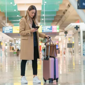 Woman looking at her smartphone to find her flight information while navigating an airport terminal with her luggage in tow.