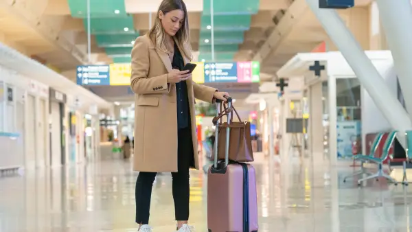 Woman looking at her smartphone to find her flight information while navigating an airport terminal with her luggage in tow.