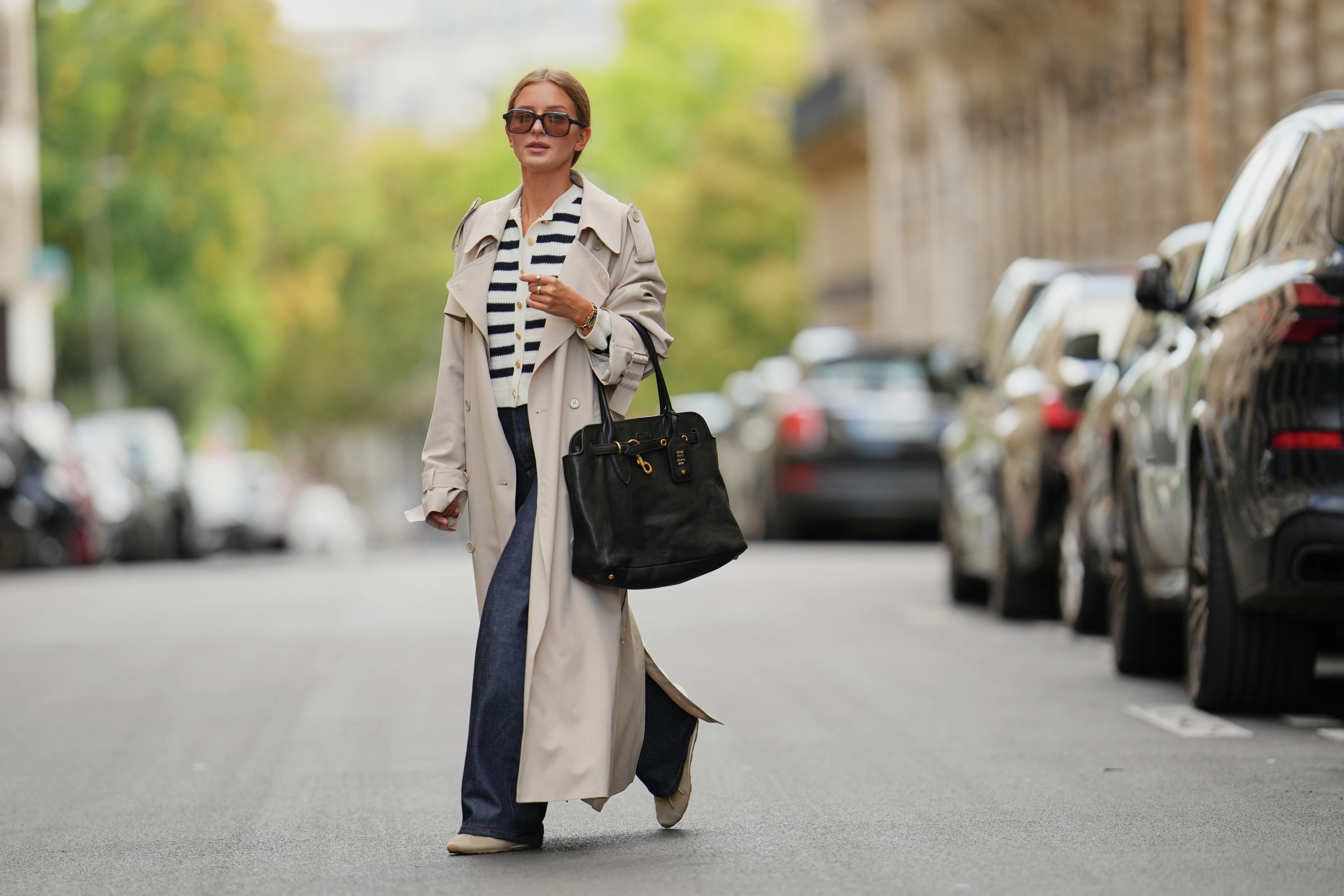 Diane Batoukina wears brown hair styled in a sleek low bun, oversized dark sunglasses by Kaleos, a beige trench coat by The Frankie Shop, a white and navy striped buttoned cardigan by APC, dark blue wide-leg jeans by APC, a black leather Miu Miu handbag with gold hardware, and beige and white sneakers by Miu Miu, during a street style fashion photo session, on September 05, 2025 in Paris, France.