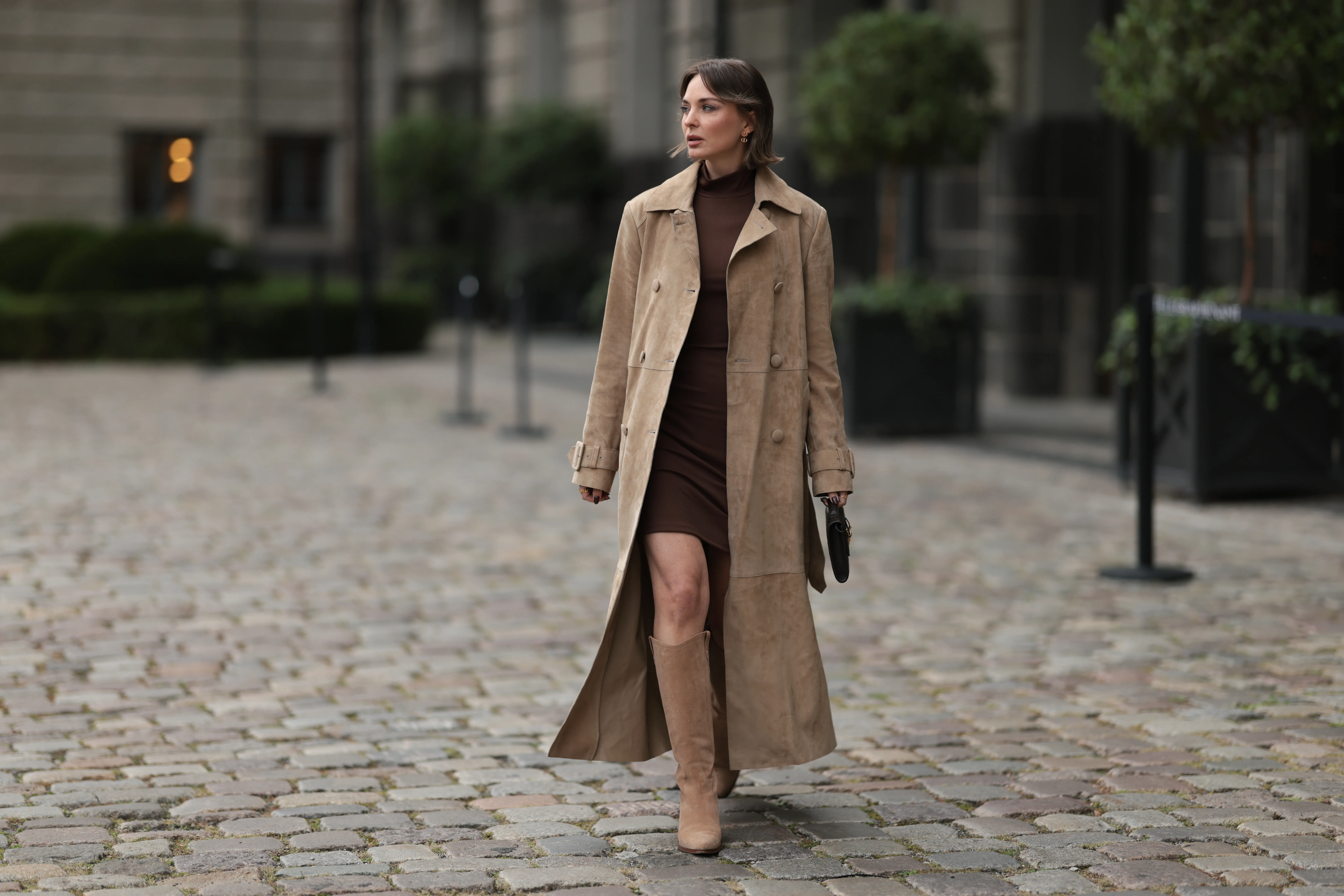 Yana Pries seen wearing a long suede leather trench coat from NOUR HAMMOUR in a warm beige tone. The coat features double-breasted buttons, a structured collar, and belted cuffs, creating a luxurious yet timeless silhouette. Her hair is cut into a sleek bob, light brown with subtle highlights, styled with a middle part and softly curved ends that frame her face. Underneath she wears Even&Odd fitted chocolate-brown mini dress, with a high neckline, Bottega Veneta Andiamo clutch, a structured black leather bag with a gold knot detail on the strap and Bobbies knee-high Bobbies suede leather boots in a soft beige tone, on September 17, 2025 in Berlin, Germany.