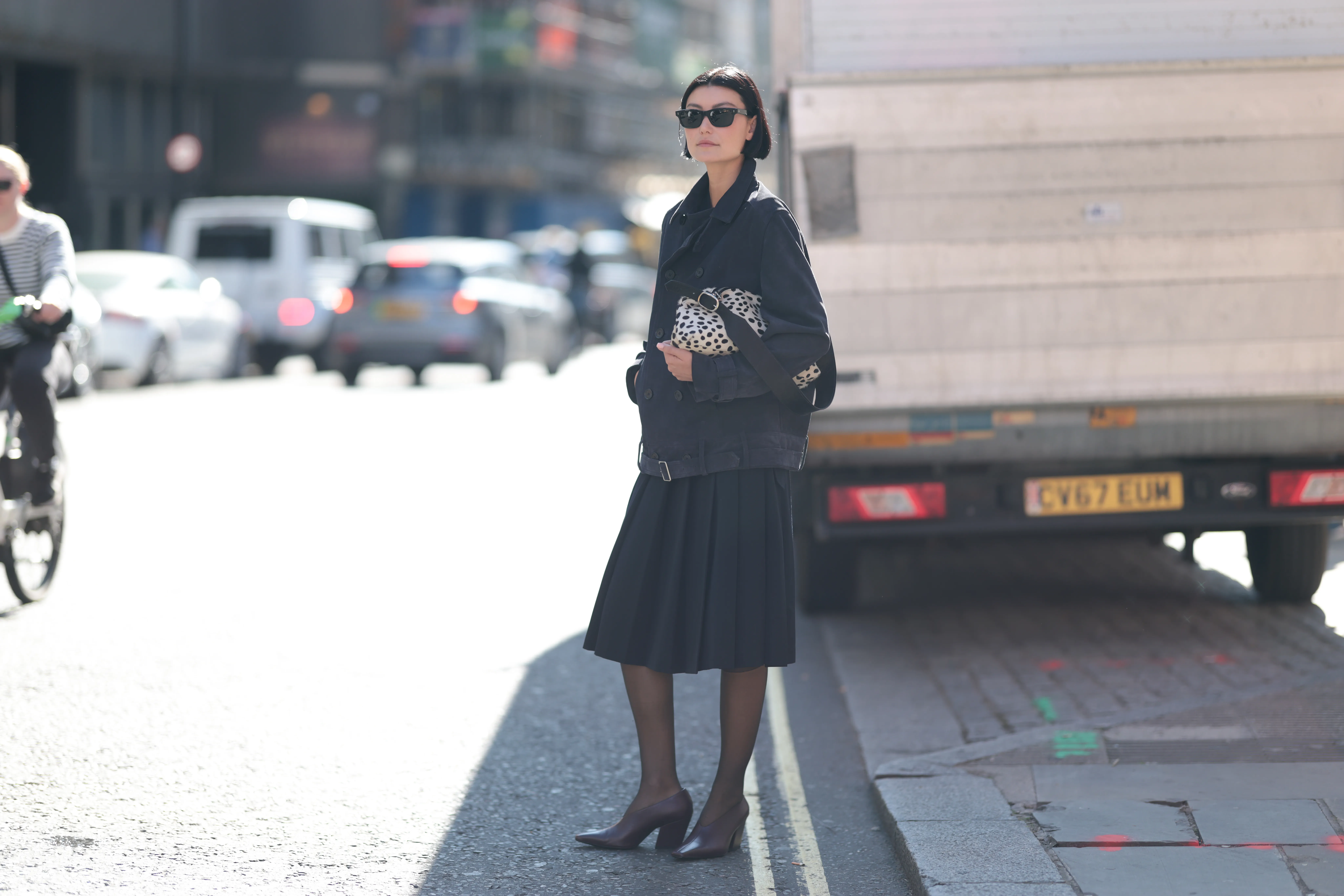 LONDON, ENGLAND - SEPTEMBER 19: Amalie Gassmann with short brown hair seen wearing all black, black short trenchcoat, pleated skirt, black tights, dark brown sandals, black sunglasses, black and white handbag with dots outside MITHRIDATE Show during London Fashion Week on September 19, 2025 in London, England. (Photo by Jeremy Moeller/Getty Images)