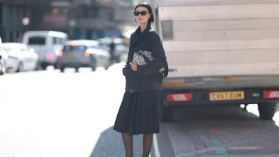 LONDON, ENGLAND - SEPTEMBER 19: Amalie Gassmann with short brown hair seen wearing all black, black short trenchcoat, pleated skirt, black tights, dark brown sandals, black sunglasses, black and white handbag with dots outside MITHRIDATE Show during London Fashion Week on September 19, 2025 in London, England. (Photo by Jeremy Moeller/Getty Images)