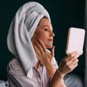 A pensive Caucasian female with her hair wrapped up in a towel keeping her face clean and hydrated using different cosmetic products. She is sitting on the bed in her bedroom. (skincare concept)