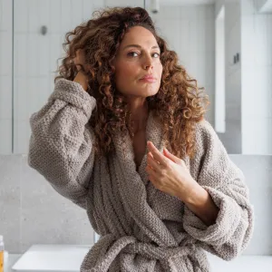 A mid-adult woman with curly hair, wearing a soft gray robe, stands thoughtfully in a bright, modern bathroom with white tiles and large windows during the daytime, fixing her hair.