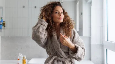 A mid-adult woman with curly hair, wearing a soft gray robe, stands thoughtfully in a bright, modern bathroom with white tiles and large windows during the daytime, fixing her hair.