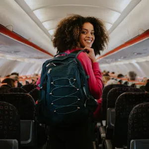 A young woman boards a small commercial airplane, carrying a backpack. She walks down the middle aisle, glances over her shoulder, and smiles at the camera.