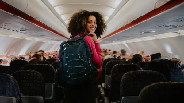 A young woman boards a small commercial airplane, carrying a backpack. She walks down the middle aisle, glances over her shoulder, and smiles at the camera.